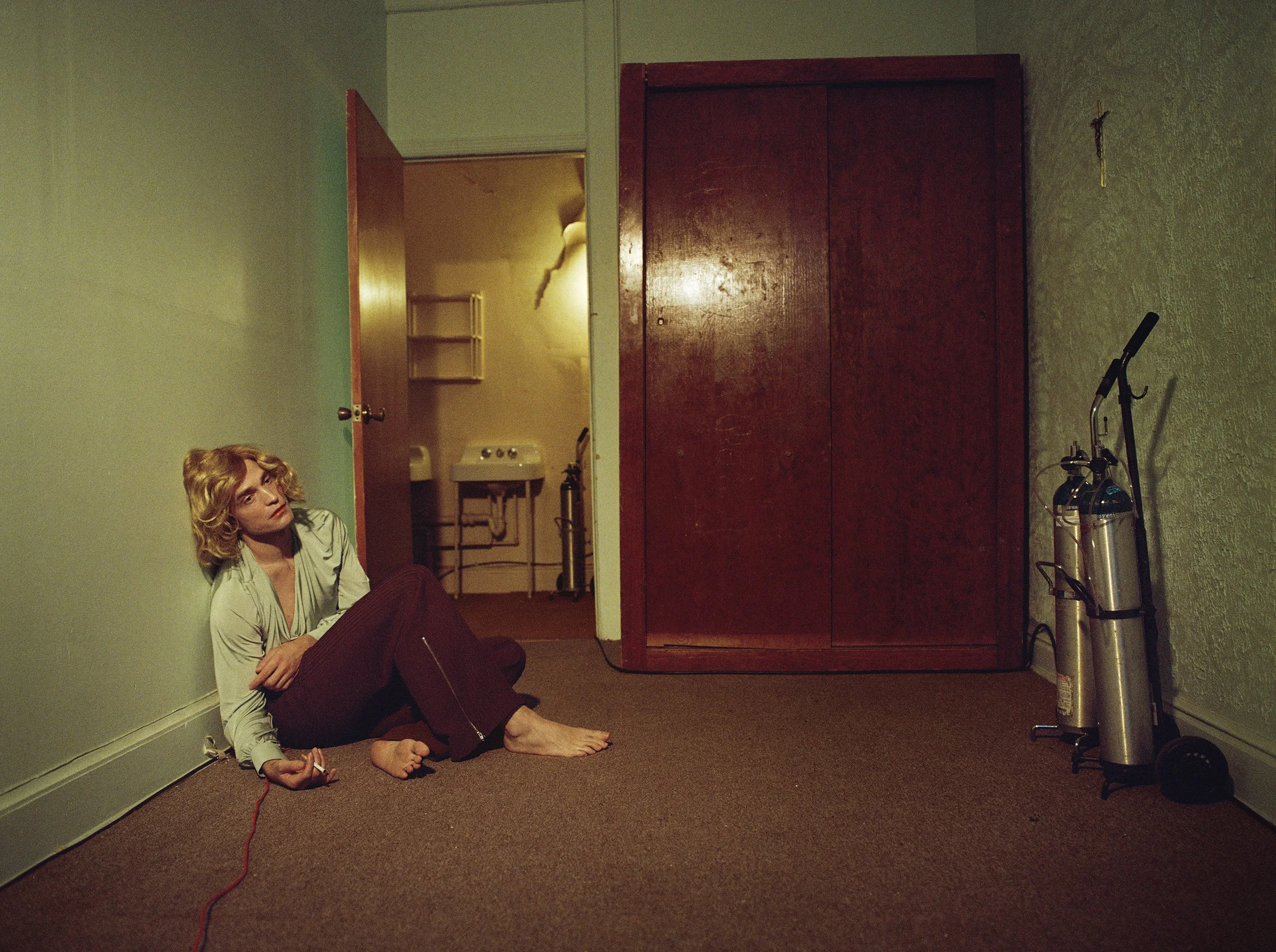 A woman with blonde hair sitting on the floor in a corner of a room, holding a plug, with an unhappy or contemplative expression. Behind her is an open door leading to a laundry room with a sink and shelves. There is a large wooden wardrobe and two f