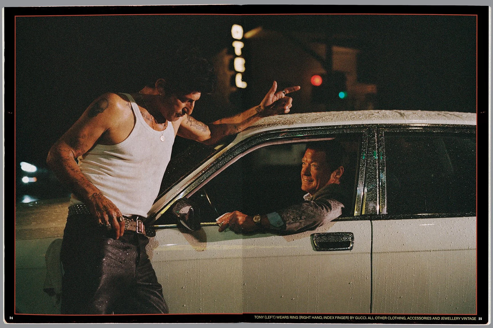 A man standing outside a car talking to a man sitting in the driver's seat during a rainy night. The man outside is wearing a white tank top and has tattoos, while the man inside is smiling and wearing a suit. The scene is illuminated by street light