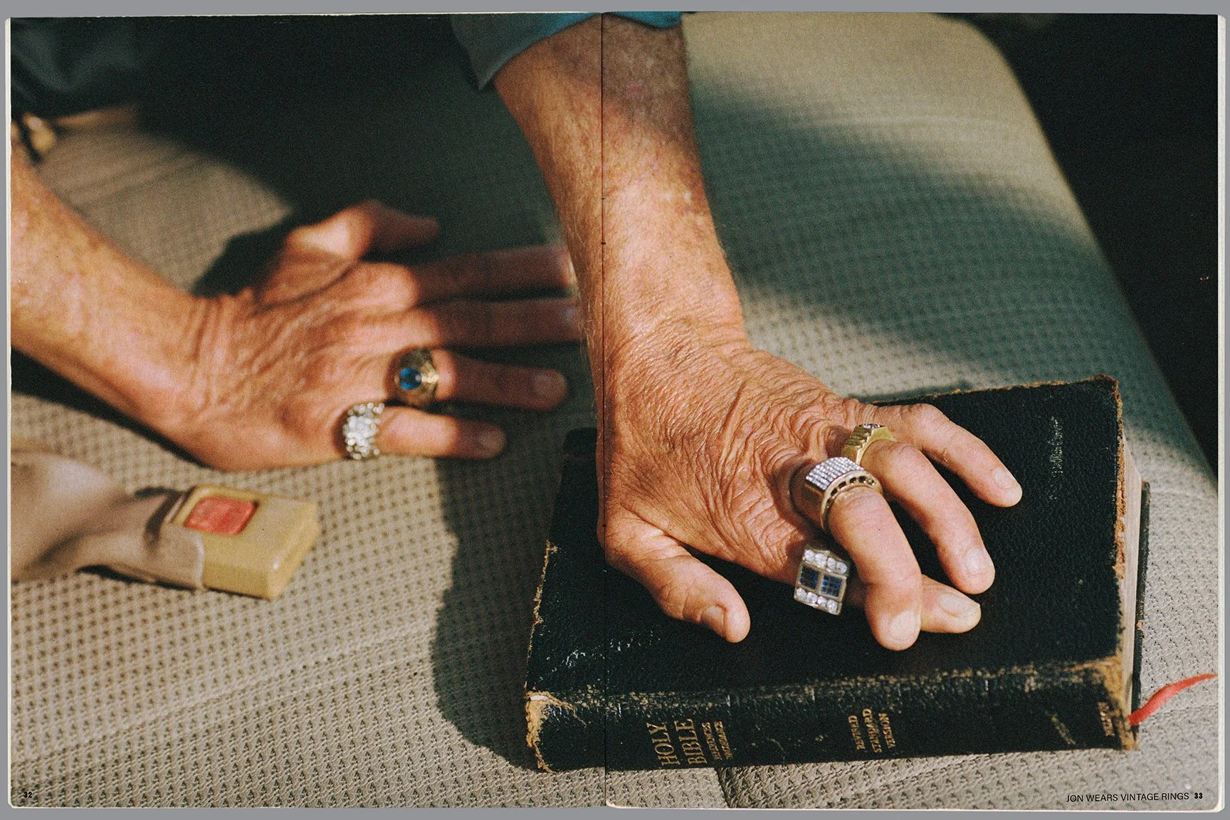 Close-up of an elderly person's hands resting on a beige fabric surface. The hands are wearing multiple vintage rings, and one hand is touching a worn black leather-bound Holy Bible. A small beige box with a red button is also on the surface next to 