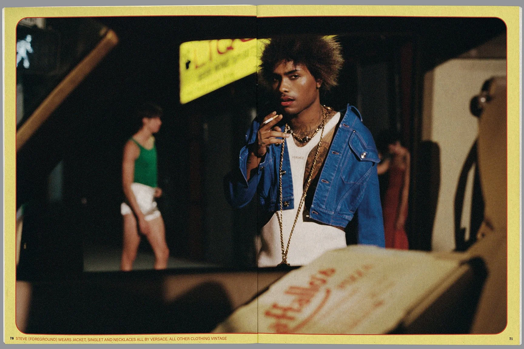 A man wearing a blue denim jacket, gold chains, and a white shirt, holding a cigarette, standing in a dimly lit environment with other people in the background, including a woman in a green top and white shorts.