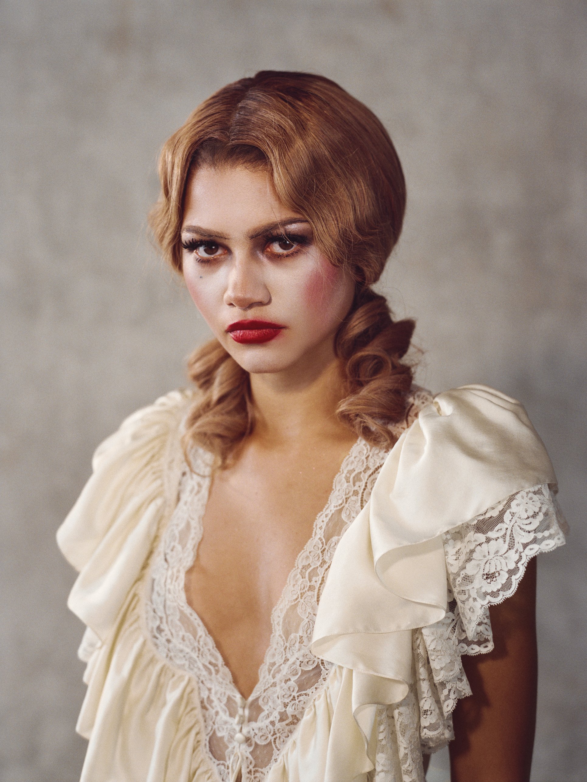 A woman with fair skin, red hair styled in waves, and bold makeup, including red lipstick and smoky eye makeup, is wearing a cream-colored dress with ruffled sleeves and lace details.