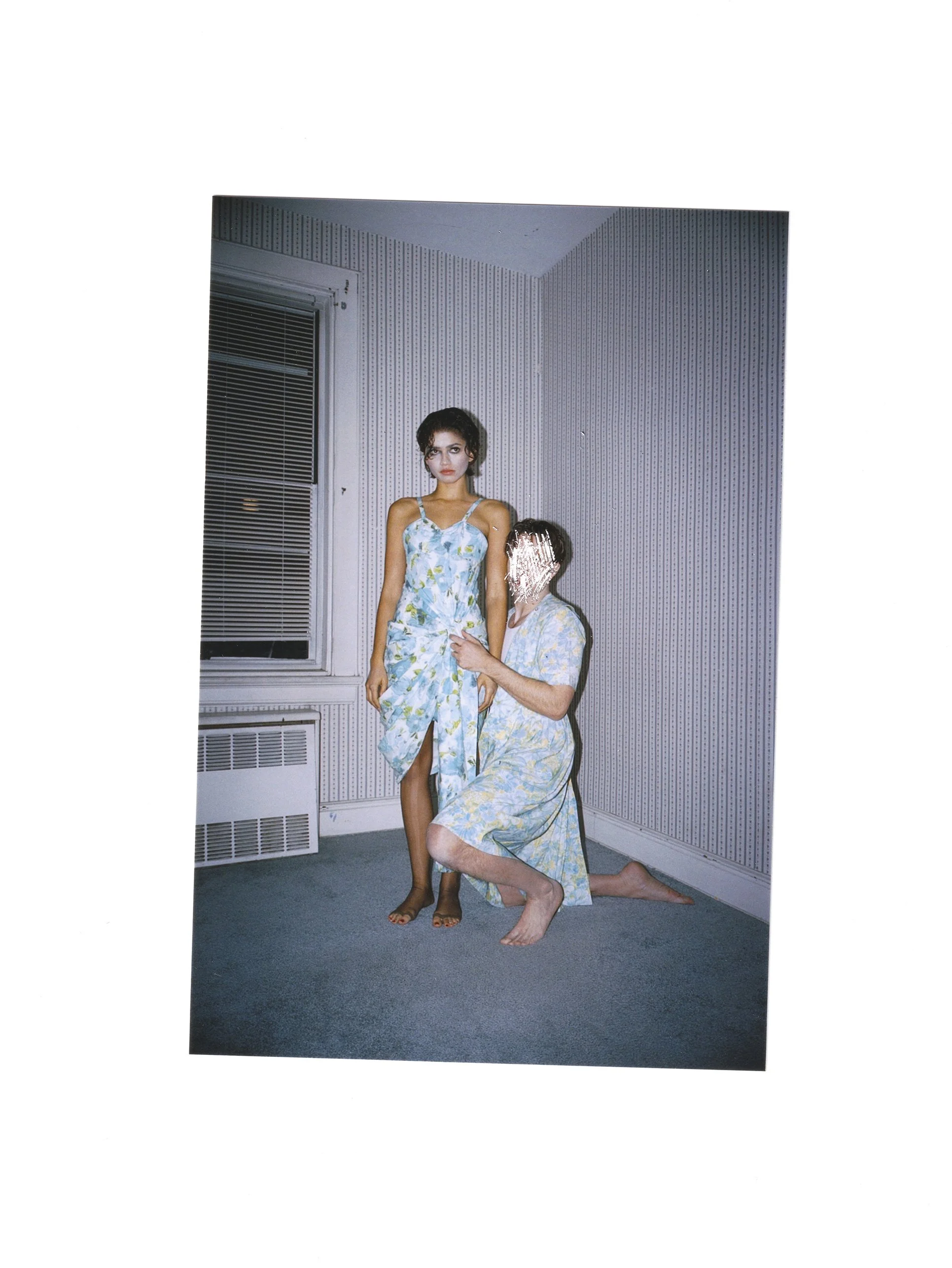 A woman standing in a room with patterned wallpaper, wearing a floral dress, with another person kneeling in front of her, possibly adjusting or adjusting her dress, and their face is obscured.