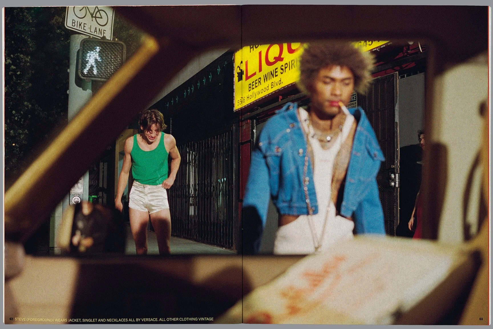 A young woman in a green tank top and white shorts is laughing on a city street at night, viewed through a broken windshield. Another person with an afro hairstyle, wearing a blue jacket and smoking a cigarette, is also visible. Street signs and a br