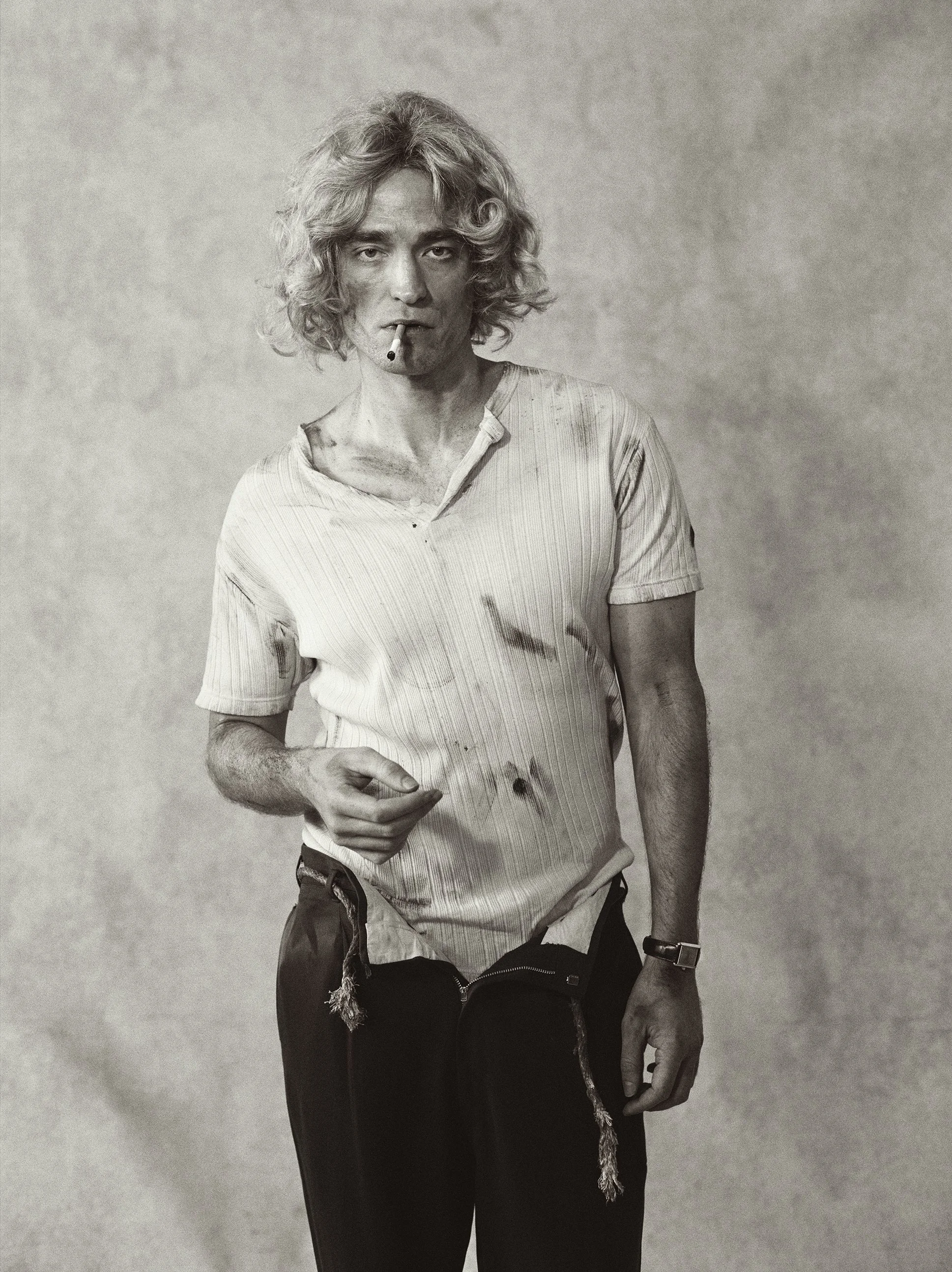Black and white photo of a man with curly hair, wearing a dirty, torn shirt and pants, holding a cigarette, with a worn and disheveled appearance.