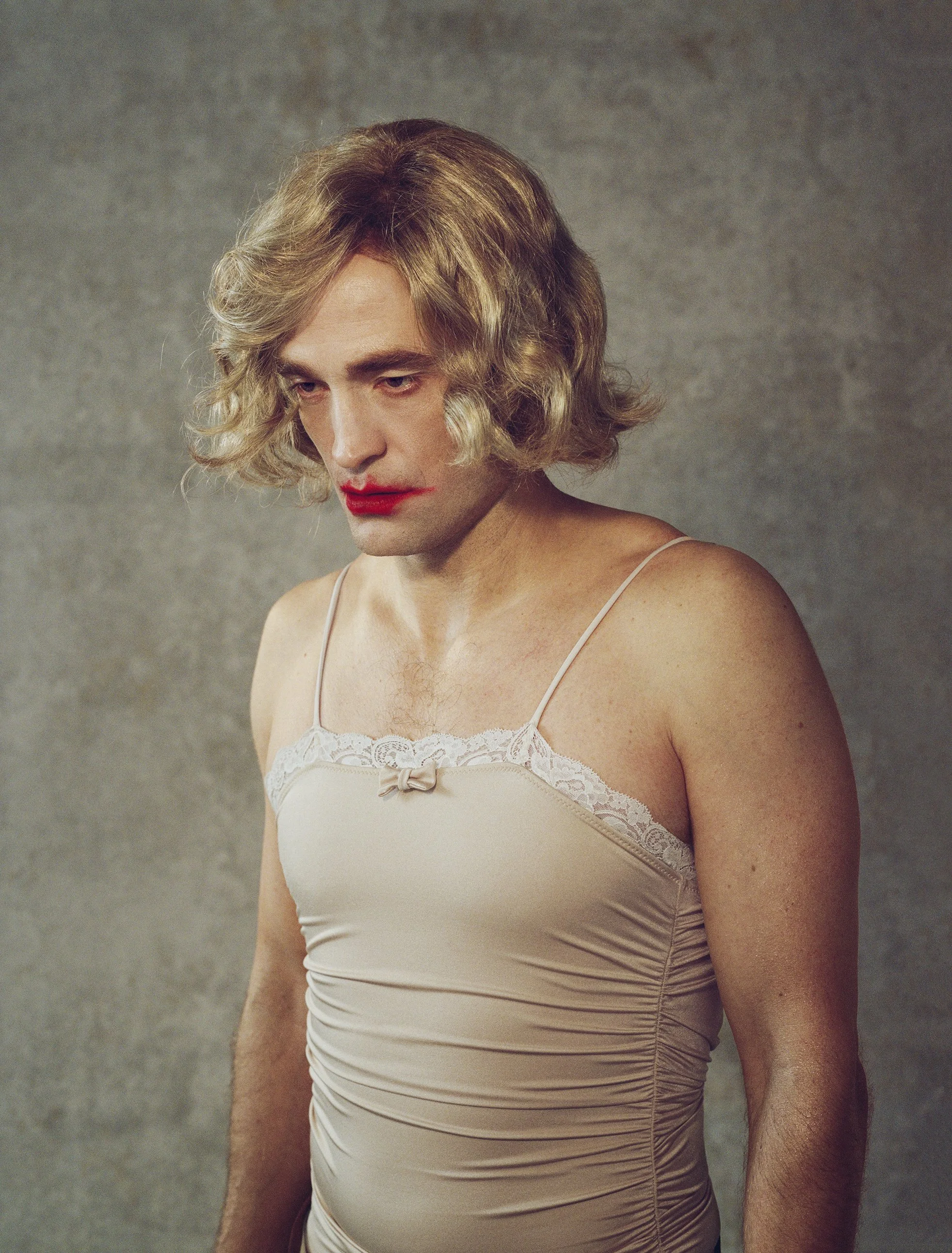 A man dressed as a woman with blonde, wavy hair, wearing a beige satin camisole with lace trim and a small bow, with red lipstick, looking downward against a plain gray background.