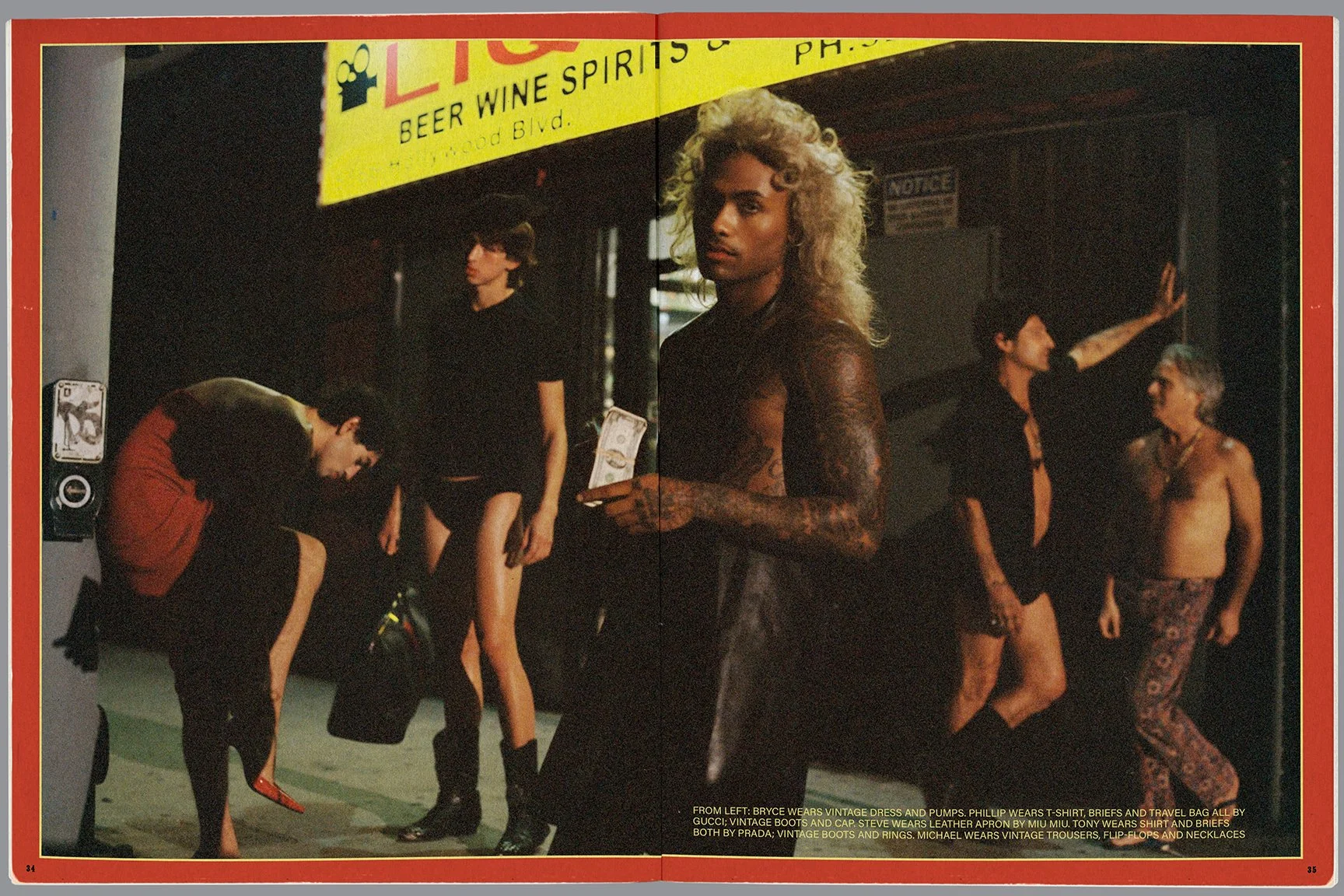 A group of fashionably dressed people standing outside a nightclub with a bright yellow sign that says 'LIQUID' and 'BEER WINE SPIRITS & PHOS'. A man with curly blonde hair, tattoos, and leather pants stands in the foreground holding a phone. To the 