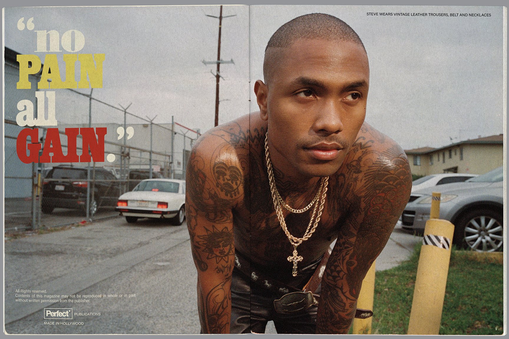 A young man with tattoos on his arms and chest, wearing multiple gold chains and vintage leather trousers, leaning forward outdoors near parked cars and yellow posts, with an overcast sky in the background.