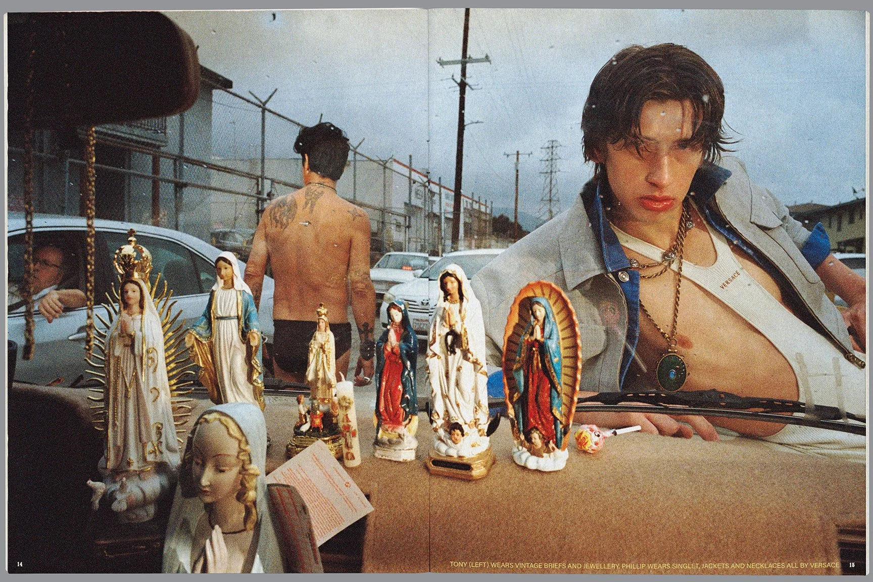 A young man with dark, wet hair wearing a partially unbuttoned vintage jacket with jewelry, looking at a display of religious figurines on a car dashboard, while a shirtless man walks away in an urban outdoor setting with cars and power lines.