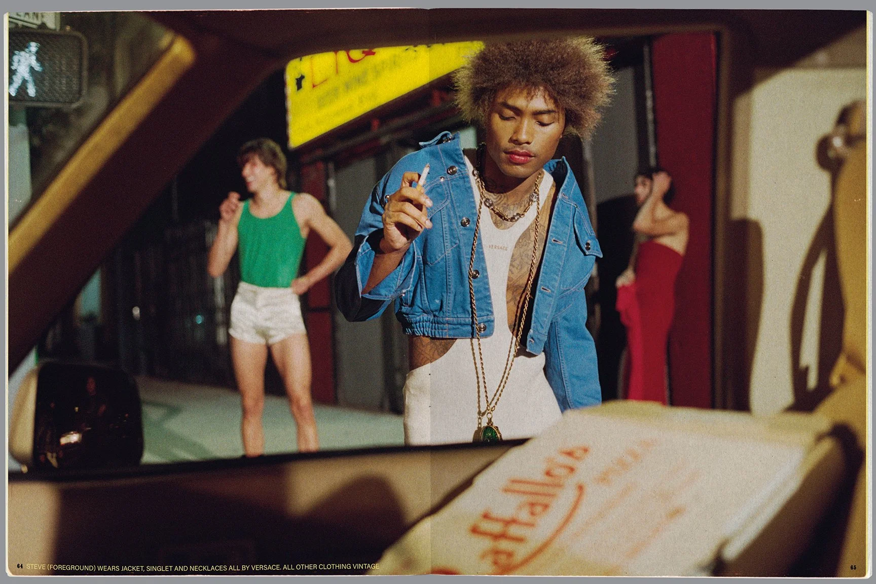 A person with curly hair is wearing a blue jacket, a white shirt, and layered gold necklaces, holding a cigarette, standing in front of a car and looking down. In the background, two women are standing on a sidewalk, one in a green tank top and white