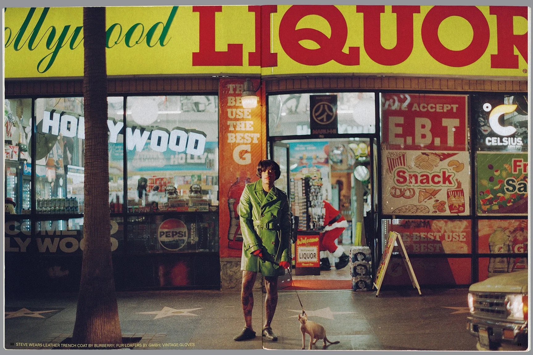 A person wearing a green leather trench coat and vintage gloves standing on a sidewalk at night, holding a leash attached to a small dog in front of a convenience store with bright red and yellow signage and a Santa Claus figure inside.