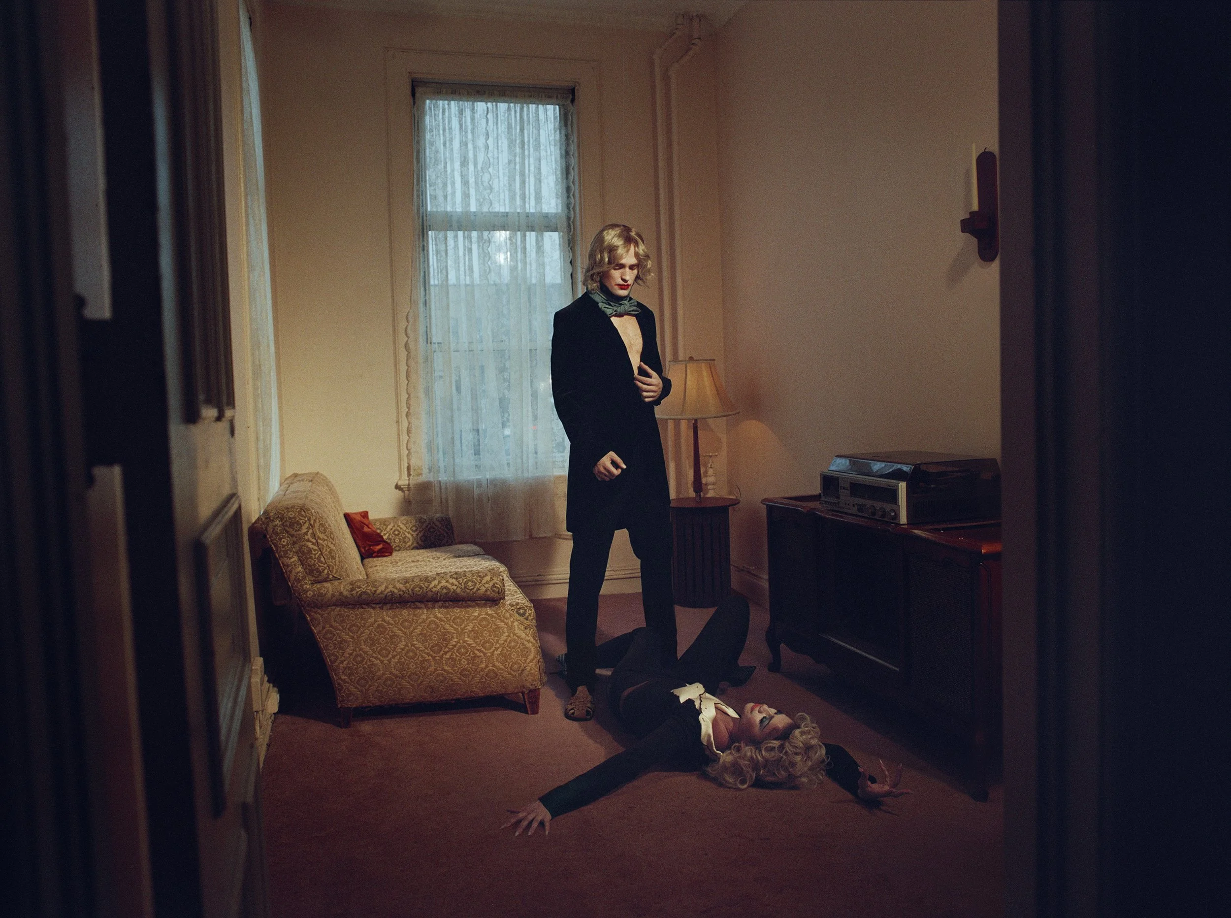A woman with blonde curly hair and vintage makeup lying on the floor with her eyes closed, arms stretched out, and wearing a black and white outfit. A woman with short blonde hair, dressed in black, stands beside her in a dimly lit vintage living roo