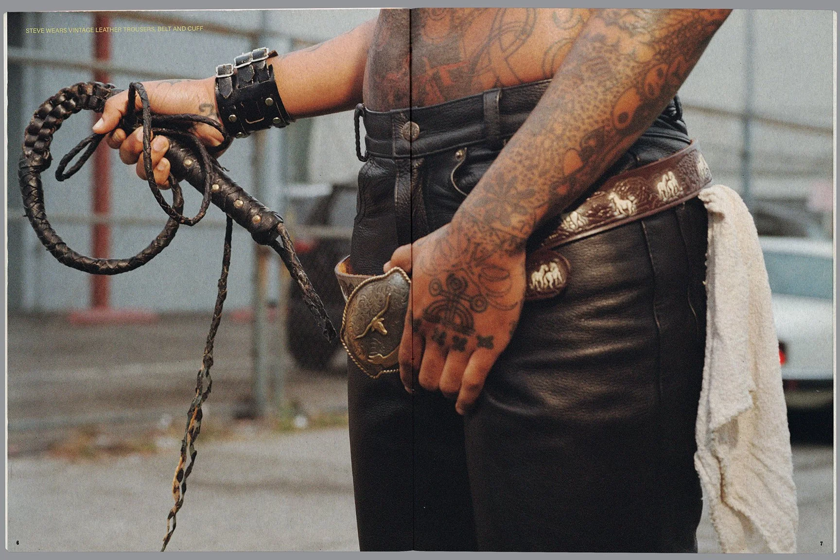 Close-up of a person's torso and hands showing tattoos, black leather pants, a belt with horse designs, and holding a leather whip, with a towel hanging from their pocket, in an urban setting.