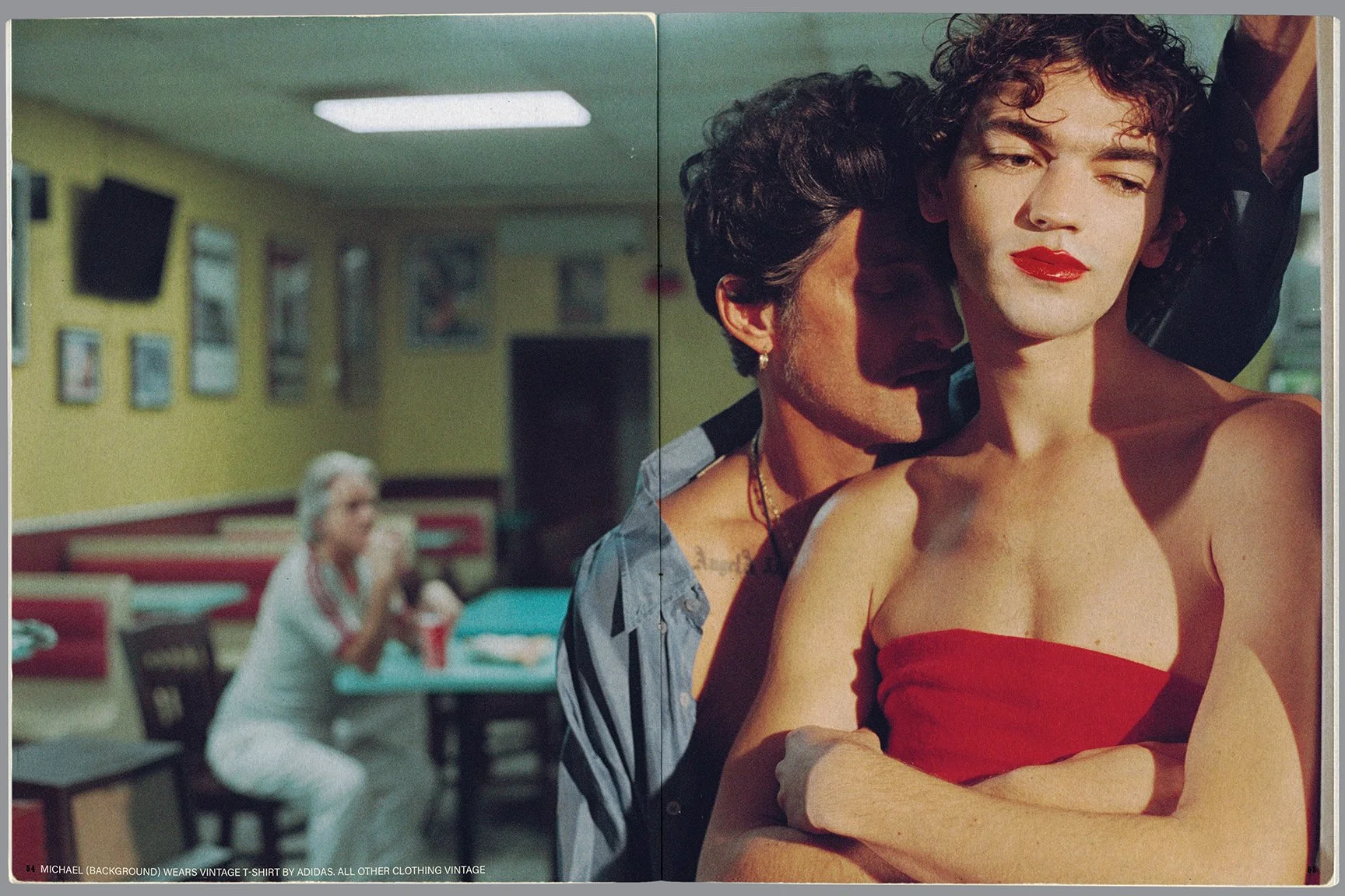 A close-up of a person with dark curly hair wearing red lipstick and a red strapless top, with another person with dark hair and earrings leaning closely behind them. In the background, an older woman is sitting at a table in a diner, wearing white c