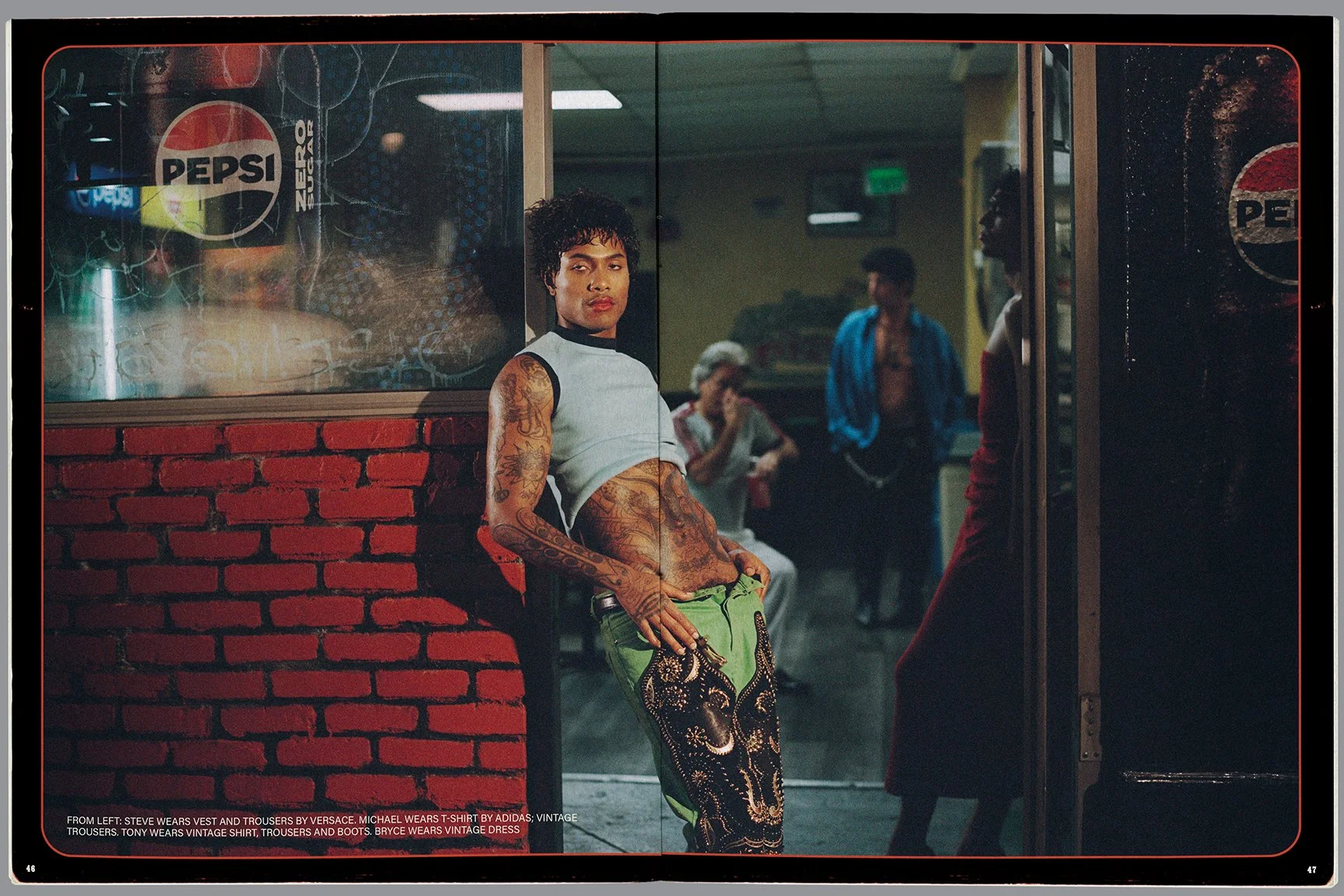 A young man with tattoos wearing a vintage vest, T-shirt, and trousers, standing outside a restaurant or shop with other people in the background.