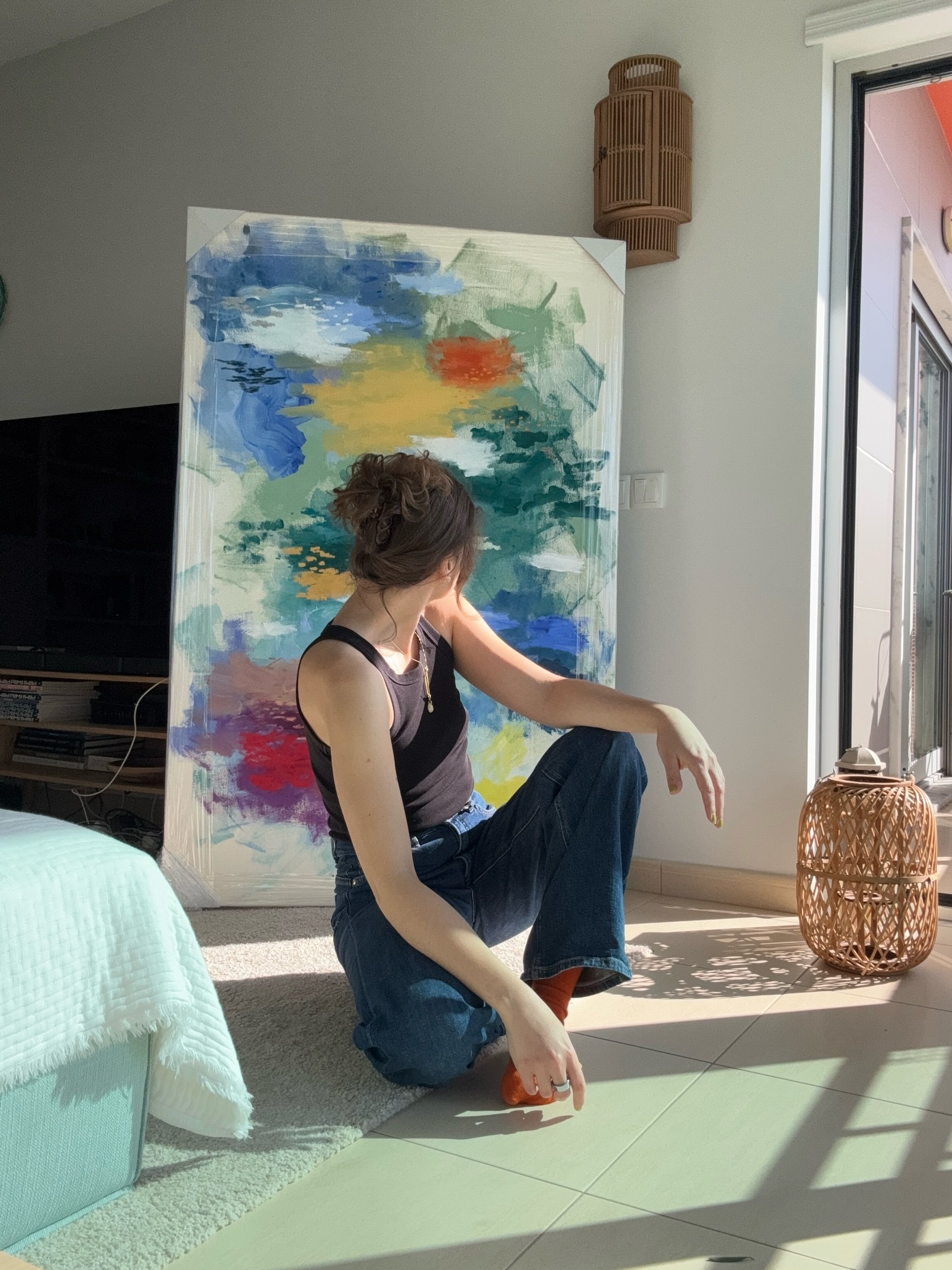 A person with curly hair wearing a black tank top and blue jeans is sitting on the floor in a brightly lit room, with one leg raised and bent at the knee. Behind them is a painted canvas with an abstract landscape in blue, green, yellow, and red. To the right, there's a wicker basket next to sliding glass doors letting in sunlight. A rattan wall lamp hangs on the wall, and a white and green blanket is draped over furniture on the left.