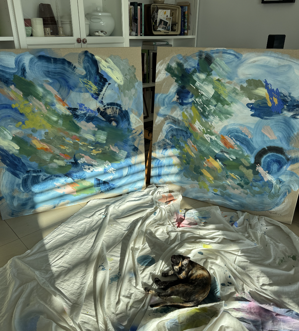 Two large abstract paintings with swirling blue, green, and yellow brushstrokes, leaning against a bookshelf in a room. A black kitten is lying on a white cloth stained with paint, in front of the paintings.