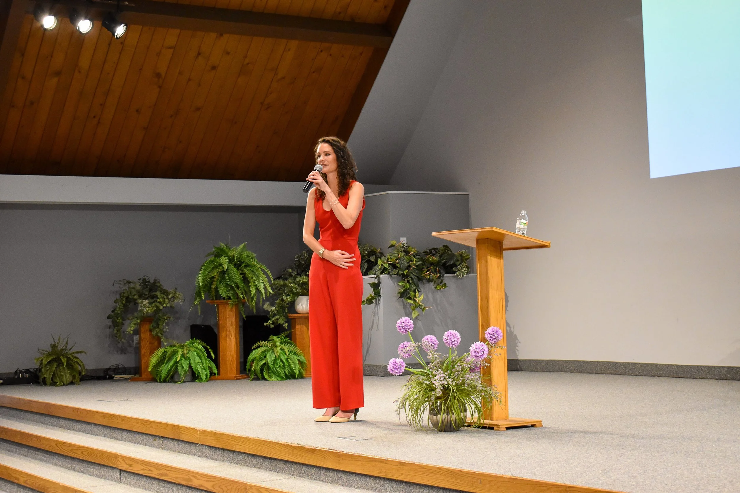A woman in a red jumpsuit stands on a stage, holding a microphone and speaking. The stage is decorated with potted plants and flowers, with a wooden podium and a large screen to the right.