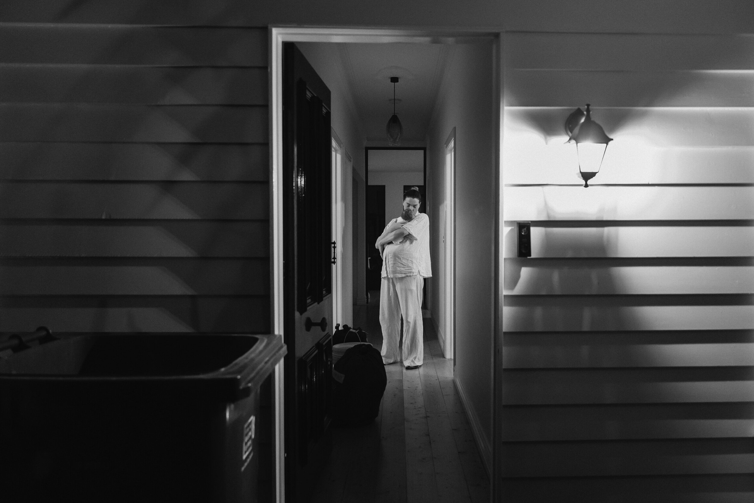 A woman in pajamas stands in a hallway clutching a cat to her chest, with a lamp hanging from the ceiling and other rooms visible in the background.