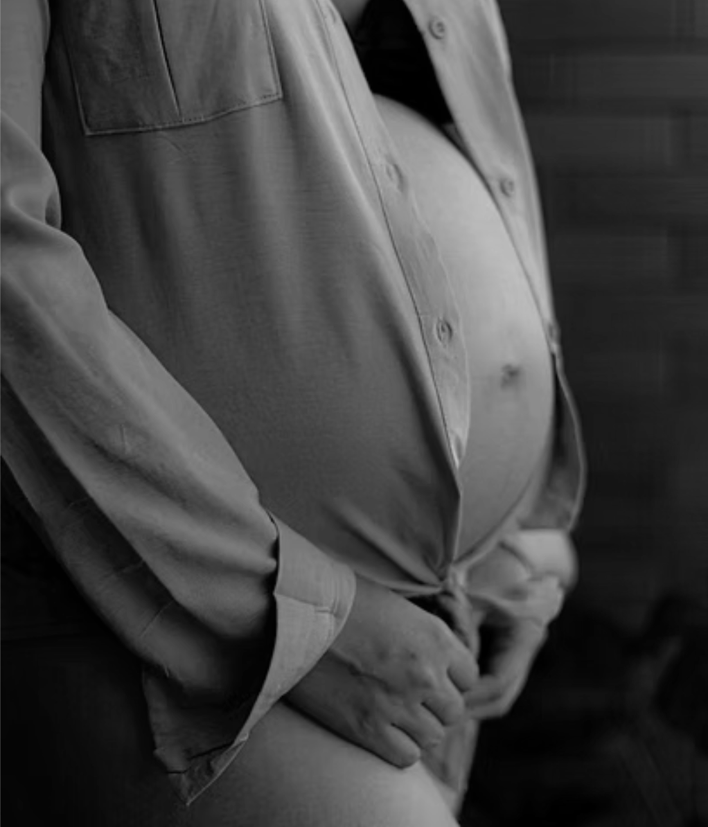 Close-up of a pregnant woman in a light-colored shirt, cradling her belly with one hand, in black and white.
