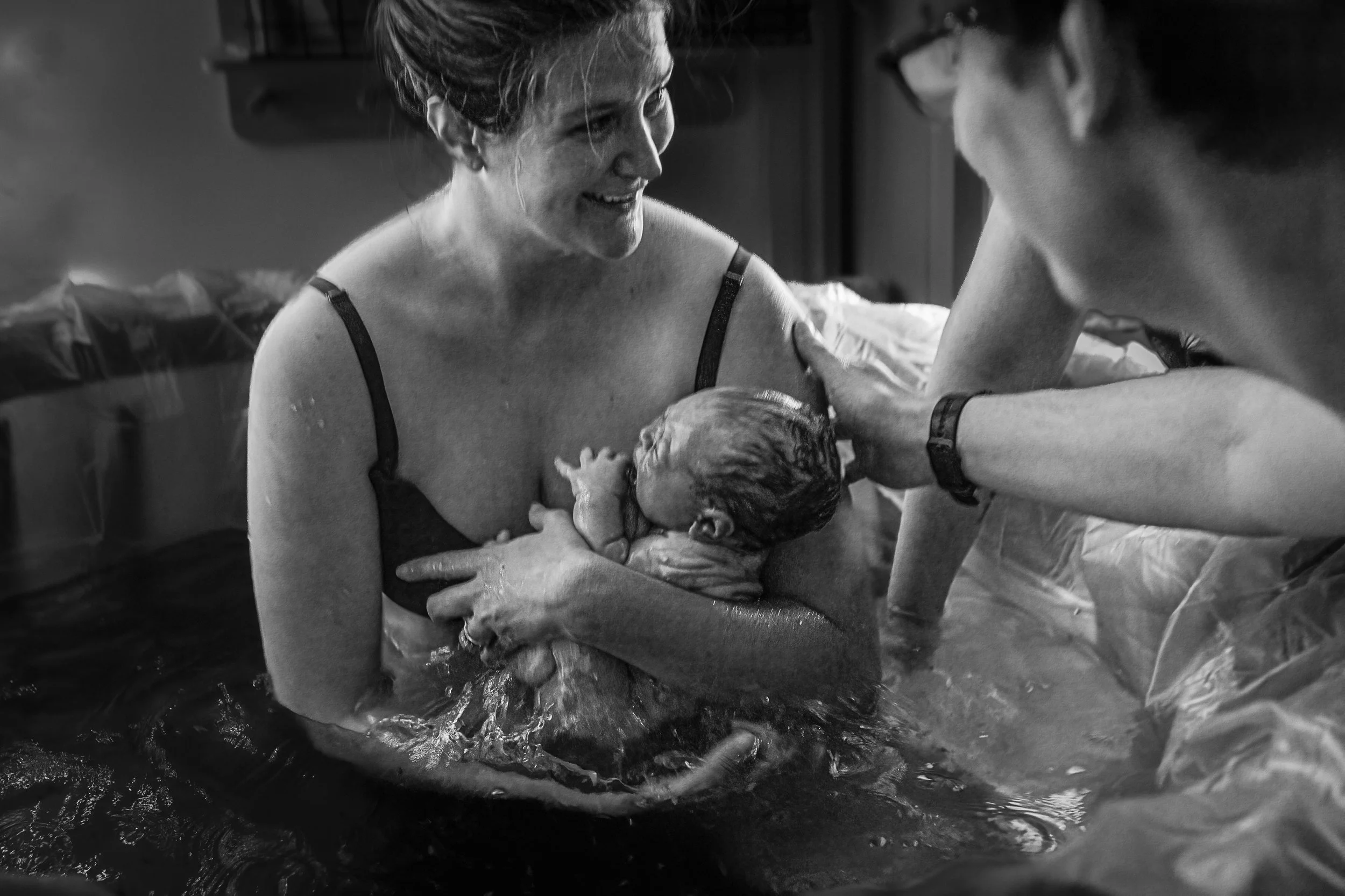 A woman is giving birth in water, holding her newborn baby, with a man touching her shoulder and an assistant supporting the baby in the water.