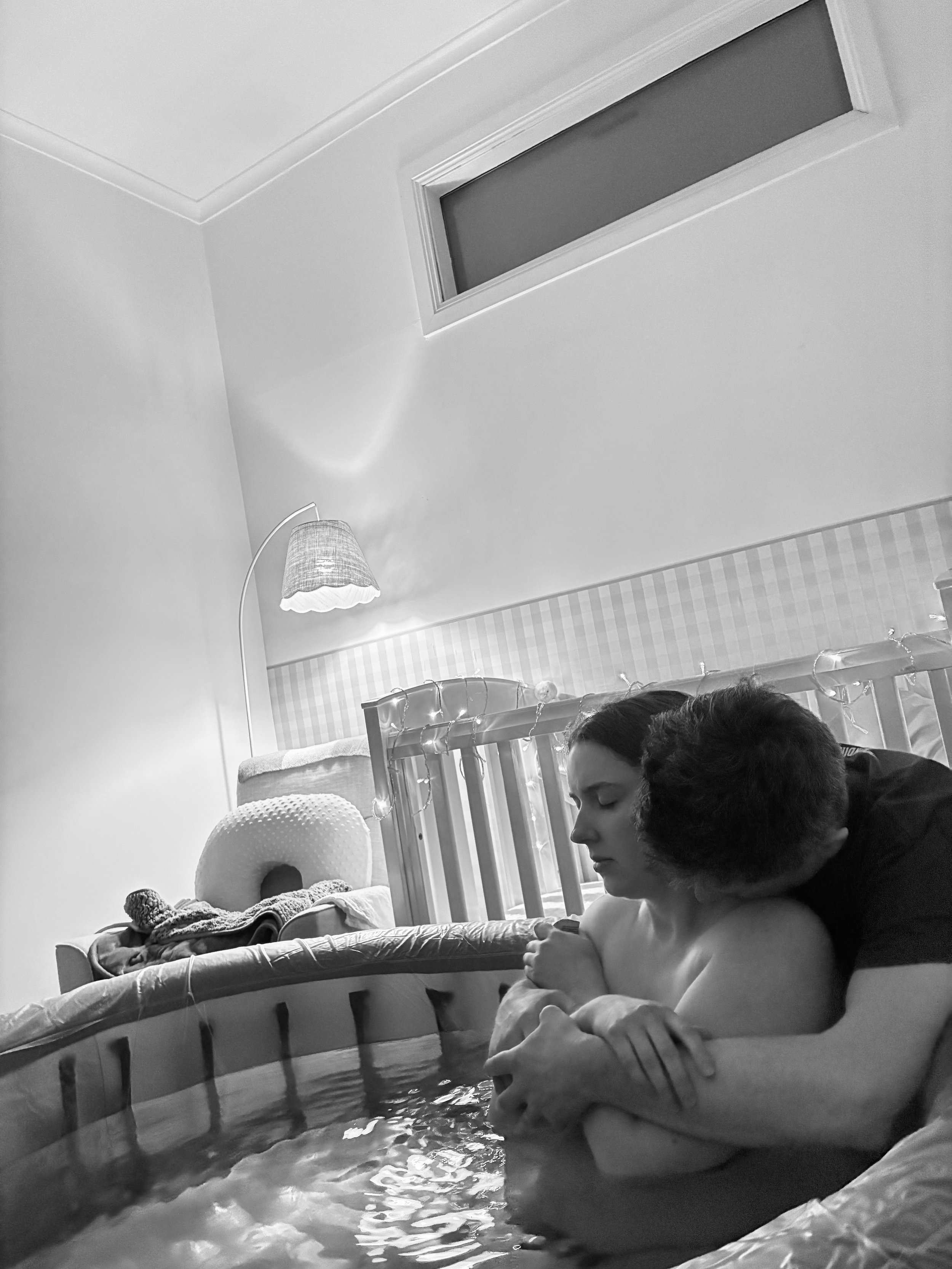 A woman and a child sitting by a small indoor pool, with the woman holding the child's arm and the child looking at the water. The room has a lit lamp, a window on the ceiling, and a bed with a headboard decorated with string lights.