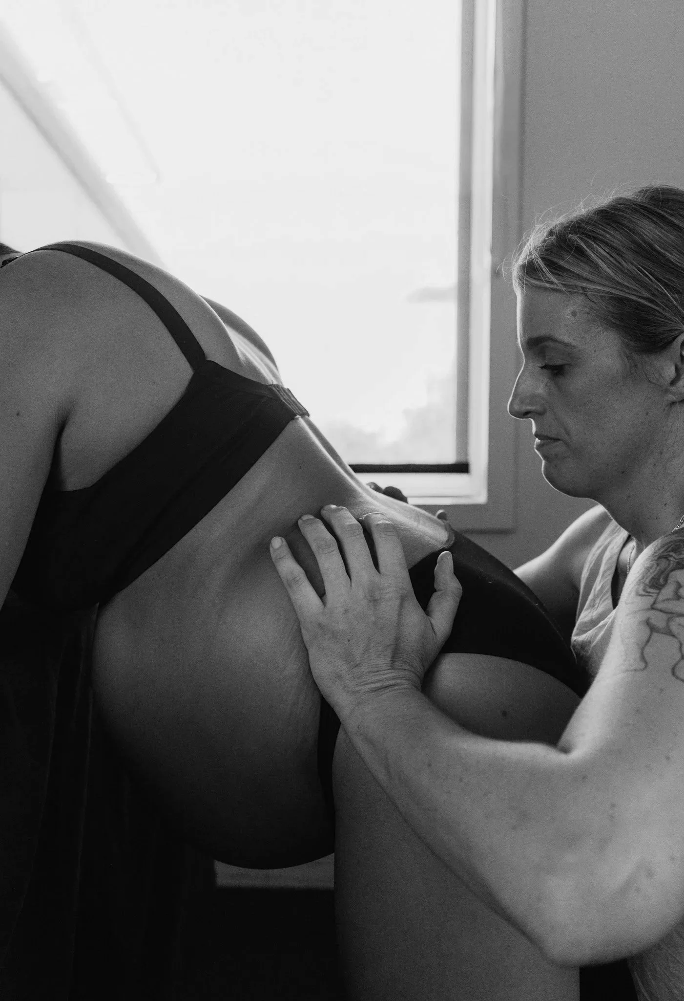 Black and white photo of a pelvic bone model placed on an anatomical diagram on a table, with a blurred background of a wall with a circular object and a framed picture.