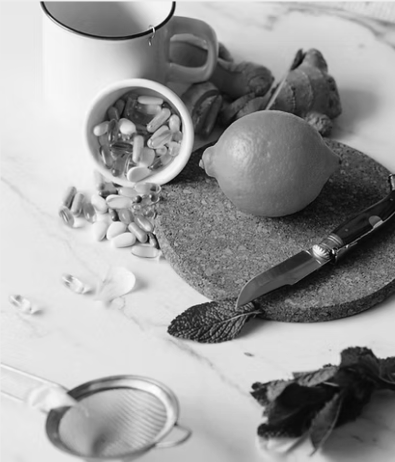A kitchen scene in black and white with a mug, an open container of capsules spilling onto a surface, a lemon, a knife, some ginger, a mint leaf, a tea infuser, and some leaves.