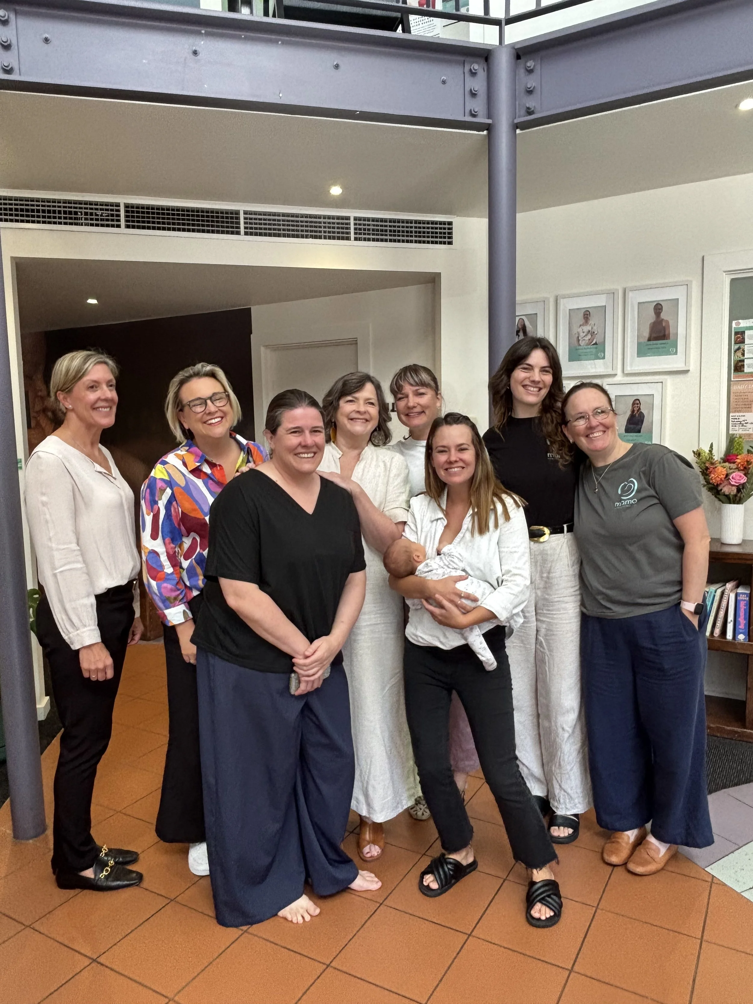 Group of women, one holding a newborn baby, smiling indoors in a casual setting.