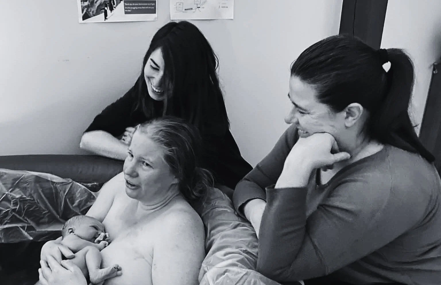 A mother holding her newborn baby after a birth pool, joined by two women, including her midwife, in a joyful moment.