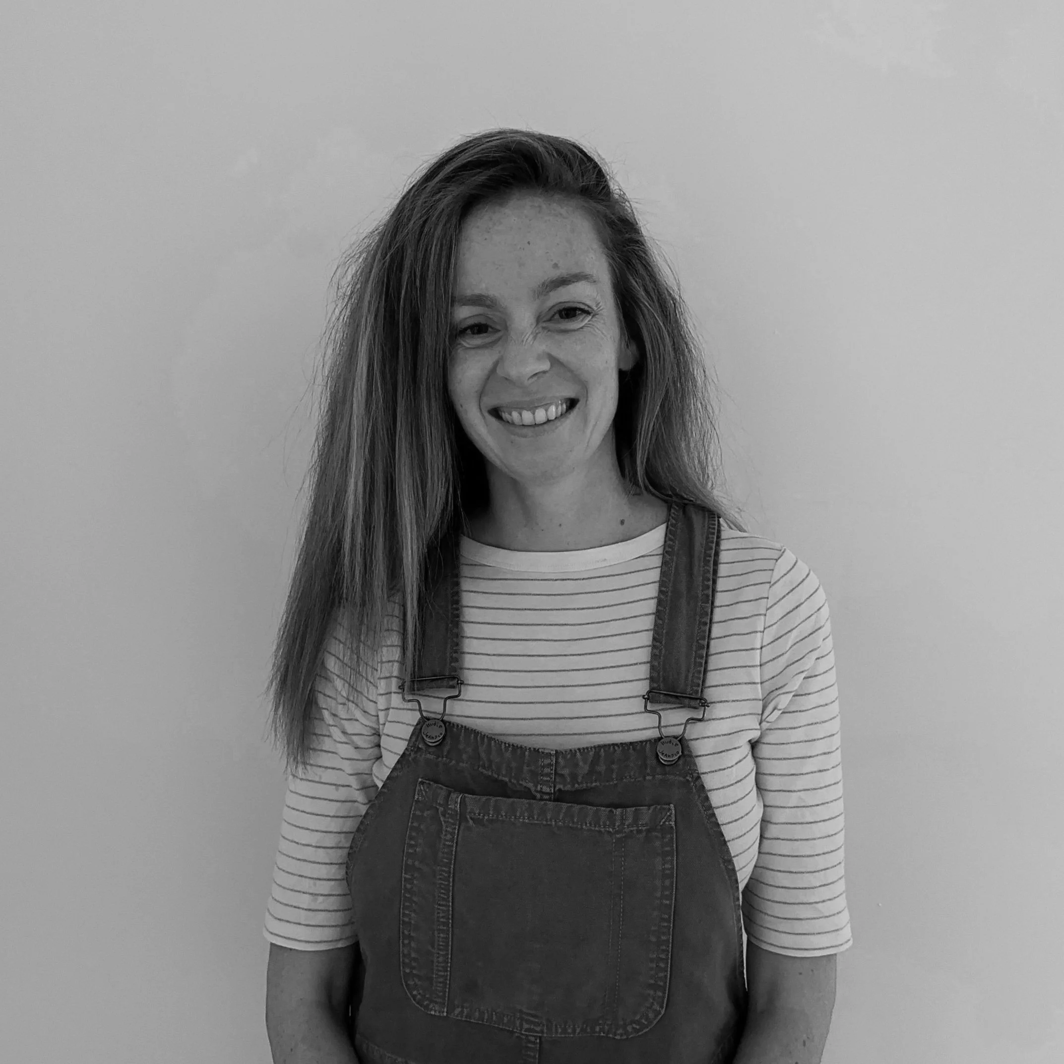 A young woman with long, wavy hair, smiling, wearing a striped long-sleeve shirt and overalls, standing against a plain background.