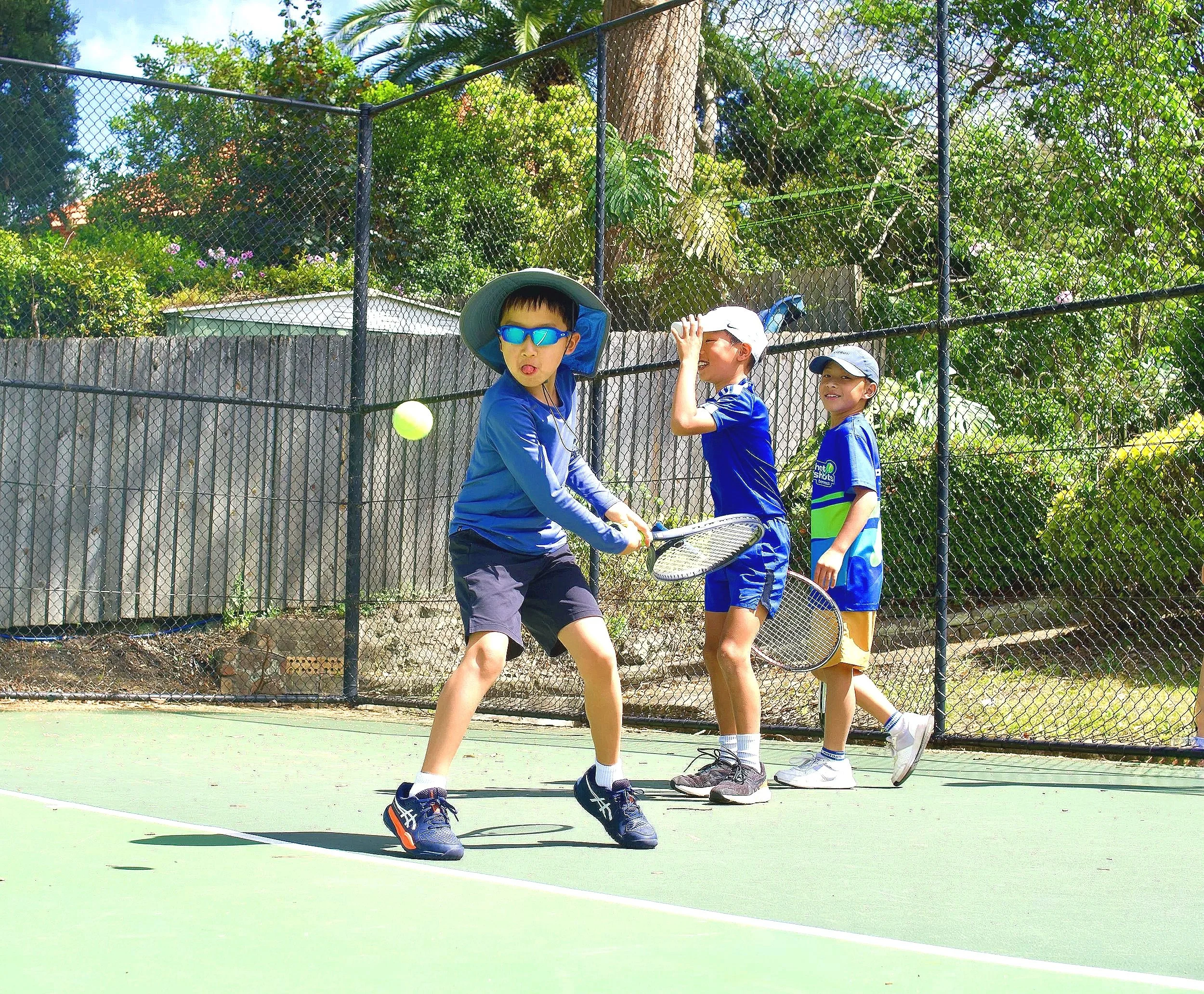 Three children playing tennis on an outdoor court, with some playing with rackets and a tennis ball, during daytime.