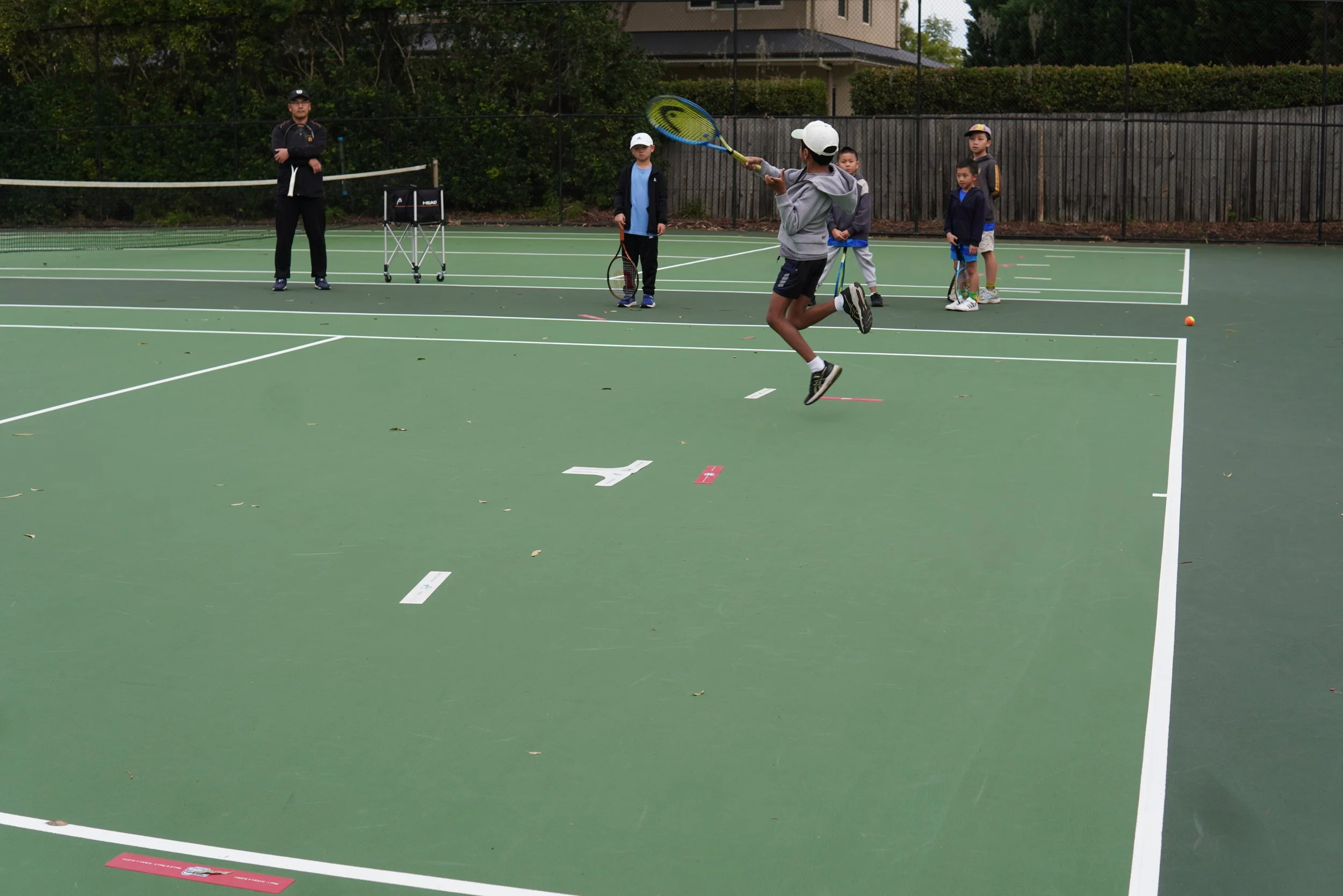 Children playing tennis on an outdoor court with an adult coach watching.