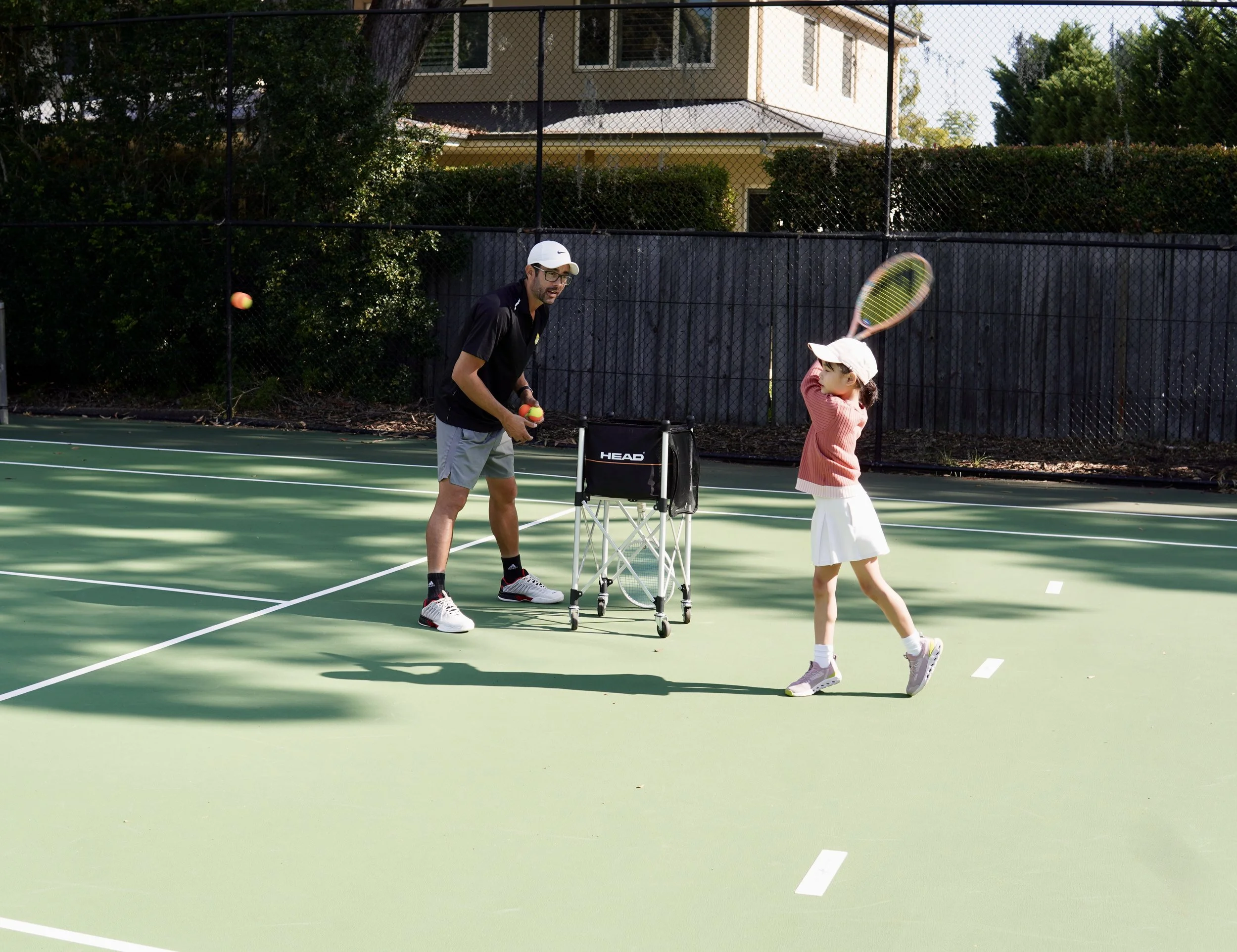 A man and a girl playing tennis on an outdoor court. The girl is preparing to hit the ball with a racket, while the man observes. Tennis balls and a ball cart are nearby.