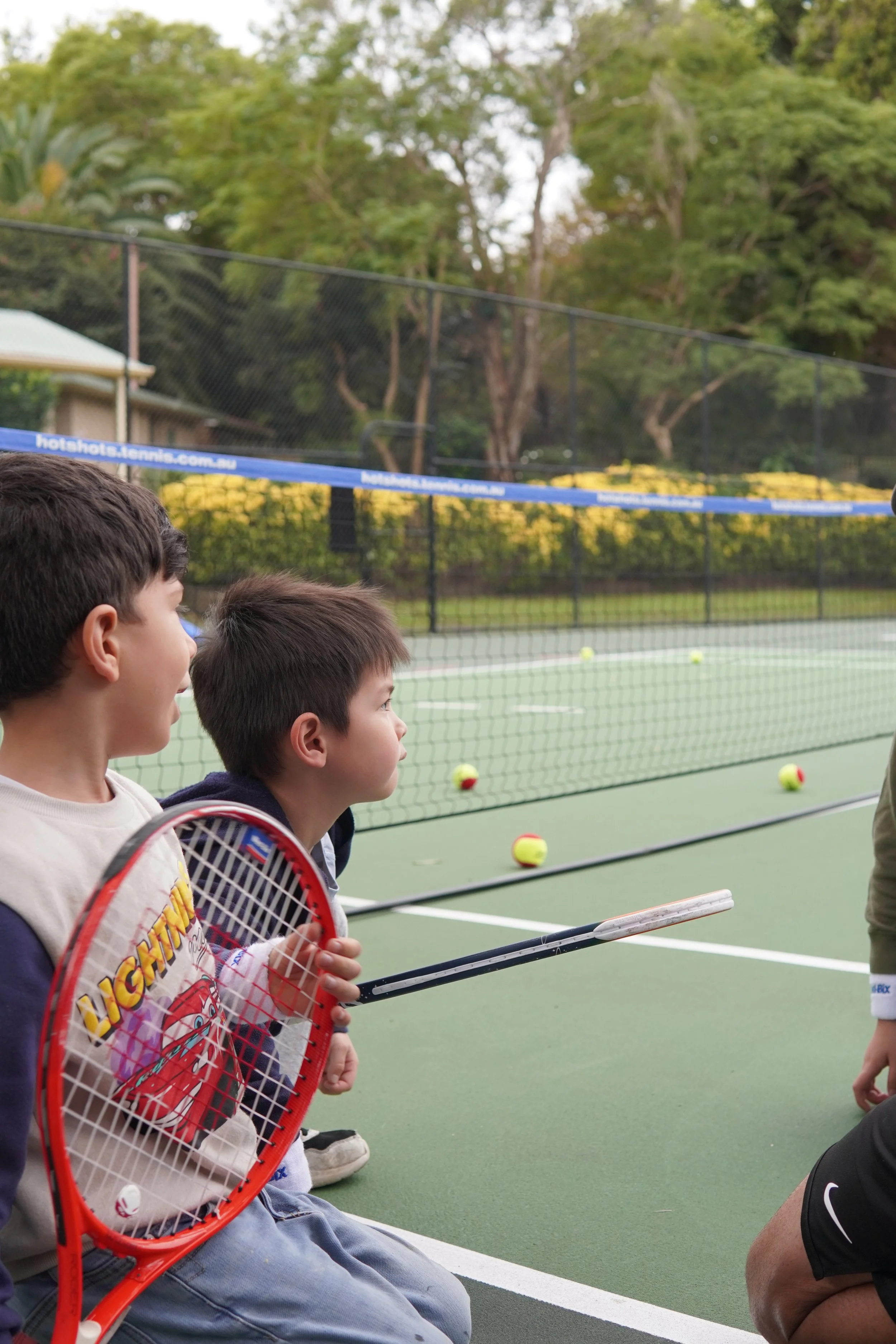 Two boys sitting on the ground and watching a tennis court with tennis balls, holding a tennis racket, during daytime.