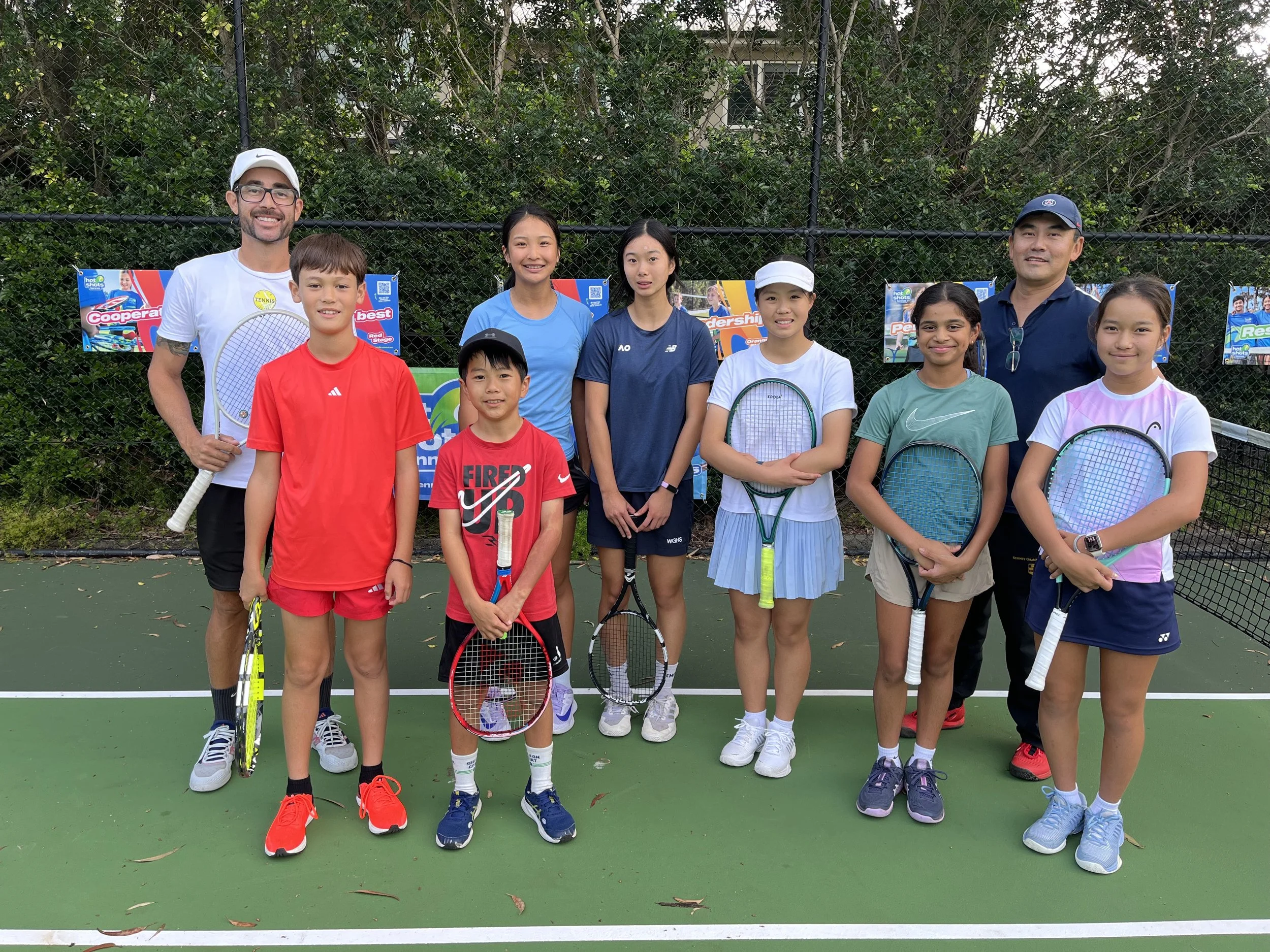 Group of eight children and two coaches standing on a tennis court after a tennis lesson, holding tennis rackets, with trees and banners in the background.