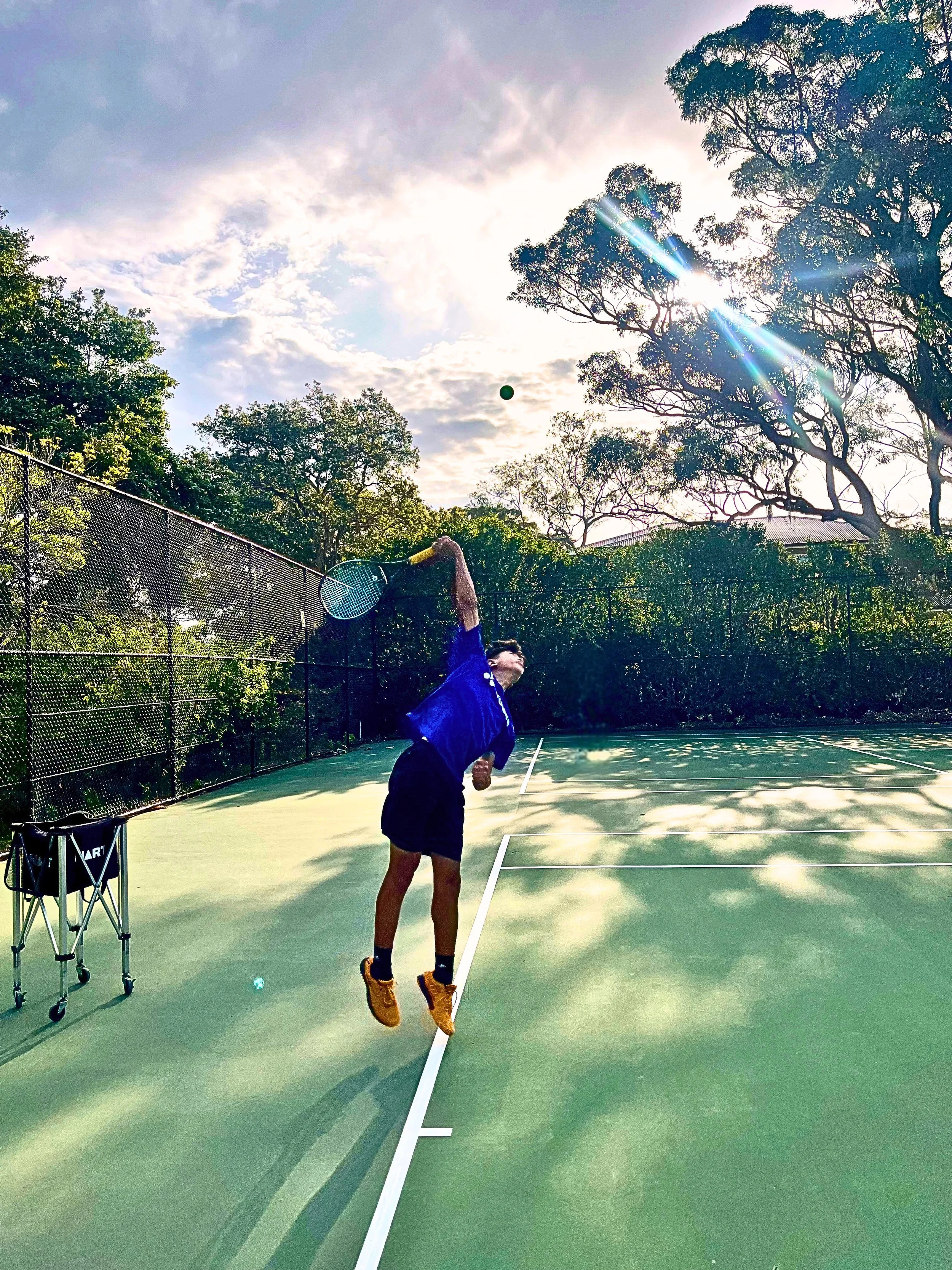 A person playing tennis on an outdoor court, about to hit a tennis ball with a racket, with trees and a partly cloudy sky in the background.