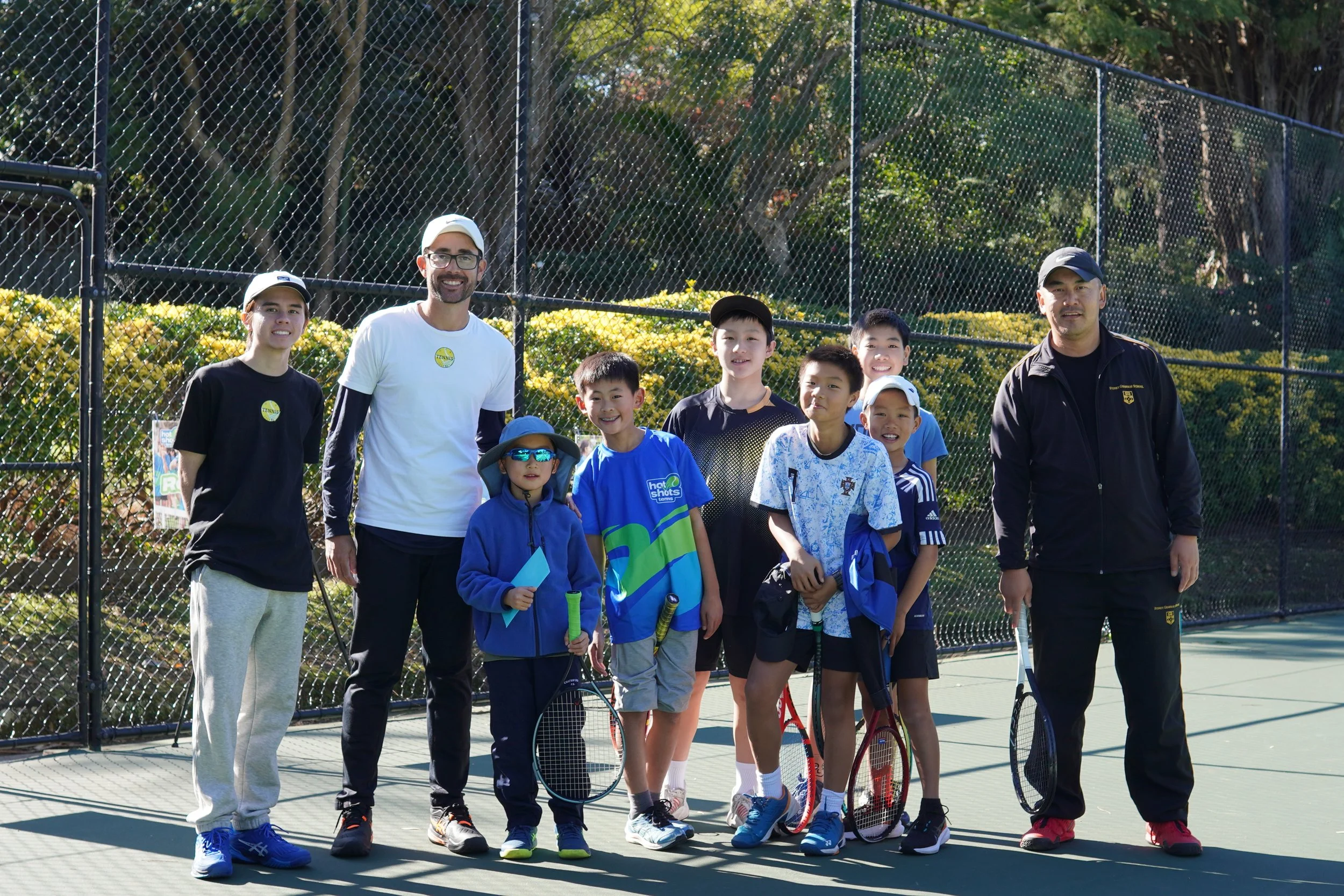 Group of young tennis players and two coaches posing on a tennis court during daytime, with a chain-link fence and trees in the background.