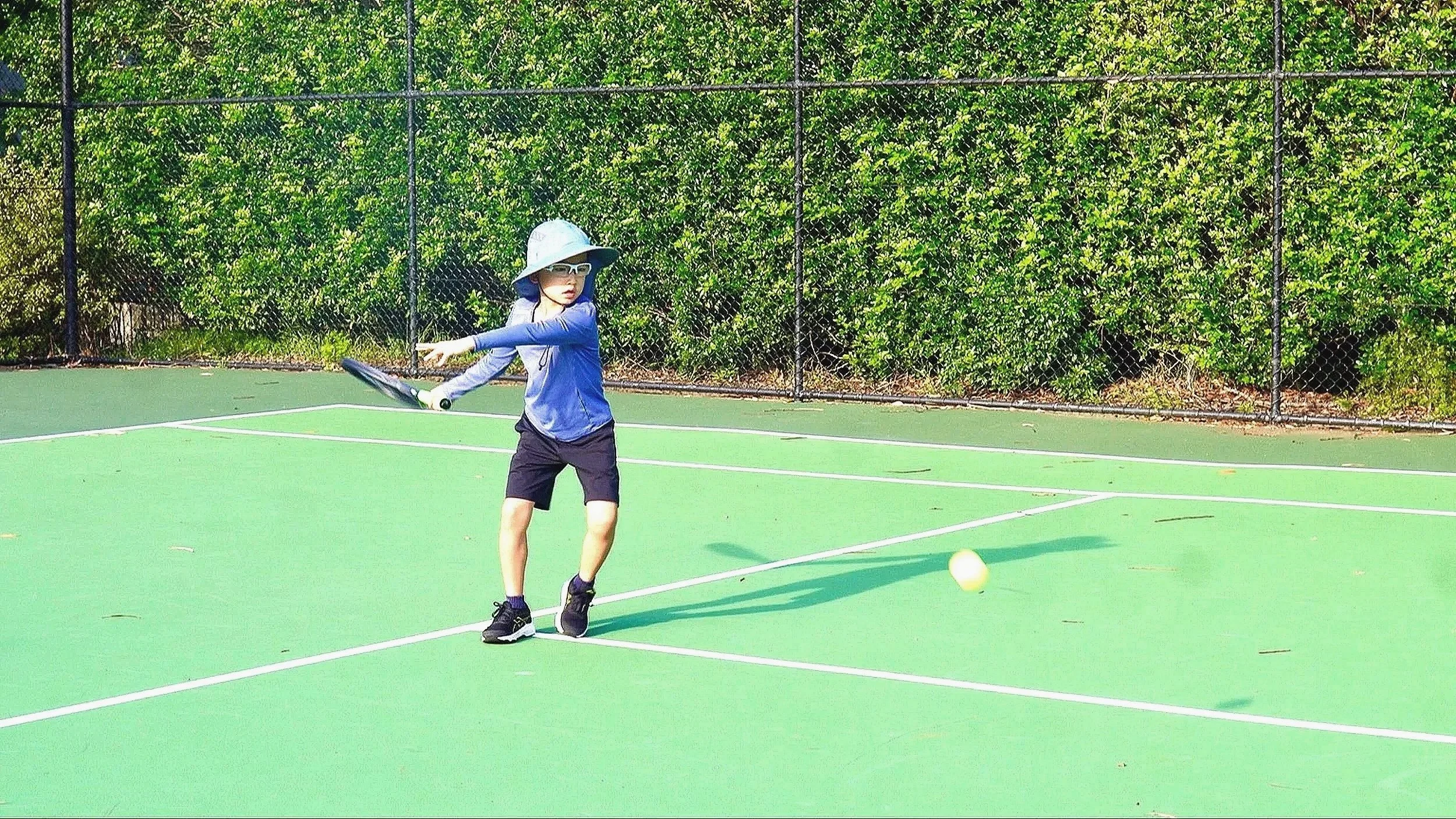 A person playing tennis on an outdoor court, jumping to hit a tennis ball with a racket in the air. The court is surrounded by trees and the sky is partly cloudy with the sun shining through.