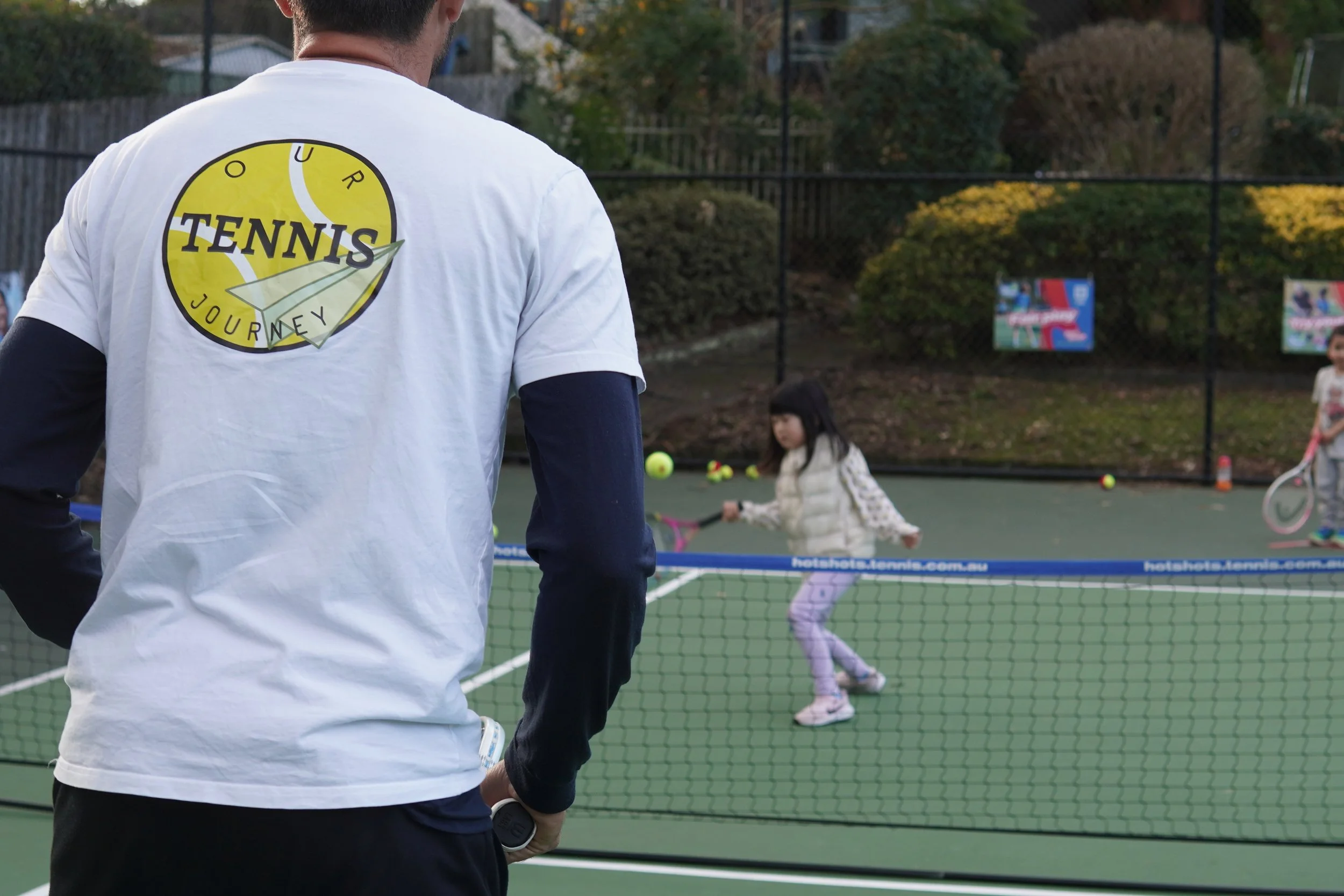 A tennis coach wearing a white shirt with a tennis logo watched children playing tennis on an outdoor court. Two girls are hitting tennis balls near the net, with one girl holding a racket. The court is surrounded by a fence and trees.