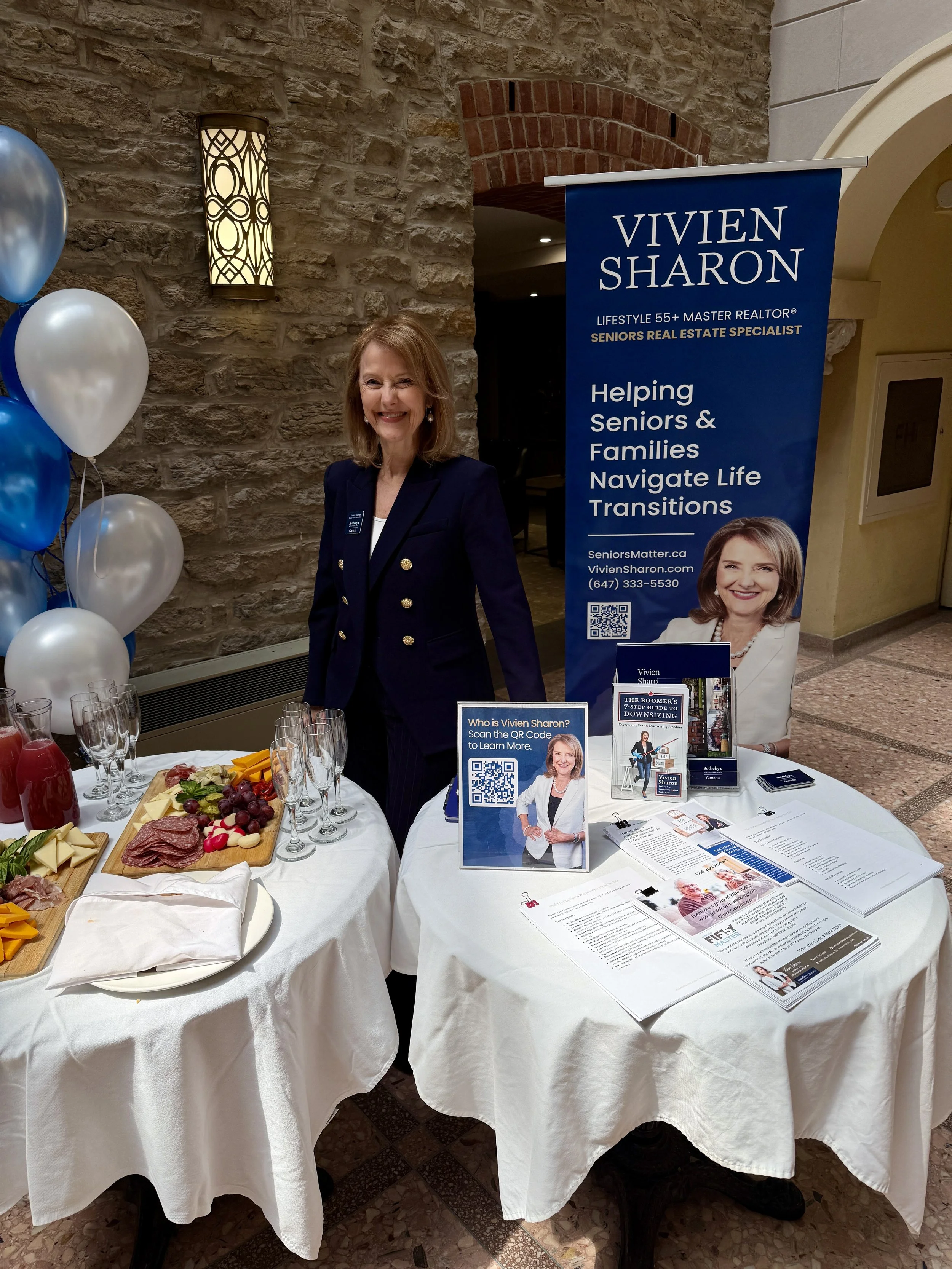 A woman at a booth for Vivien Sharon, a senior real estate specialist, standing beside a table with brochures and food, in a building with stone walls and balloons.