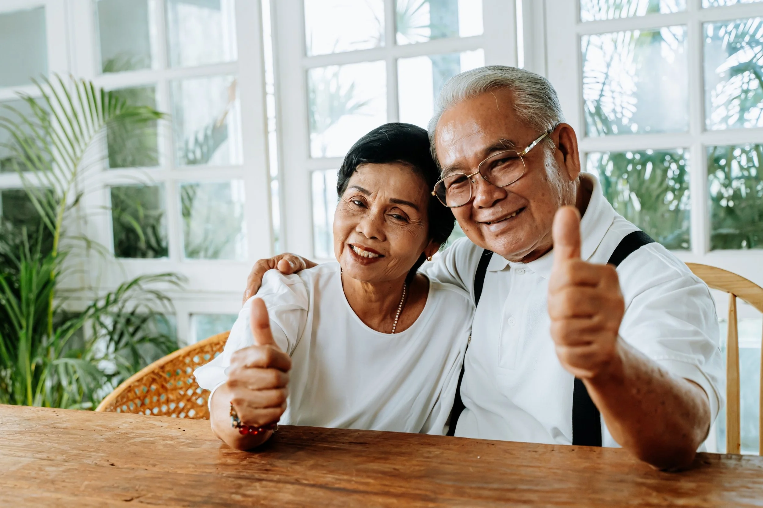 An elderly couple sitting at a wooden table, smiling, giving thumbs up, in a bright room with large windows and green plants in the background.