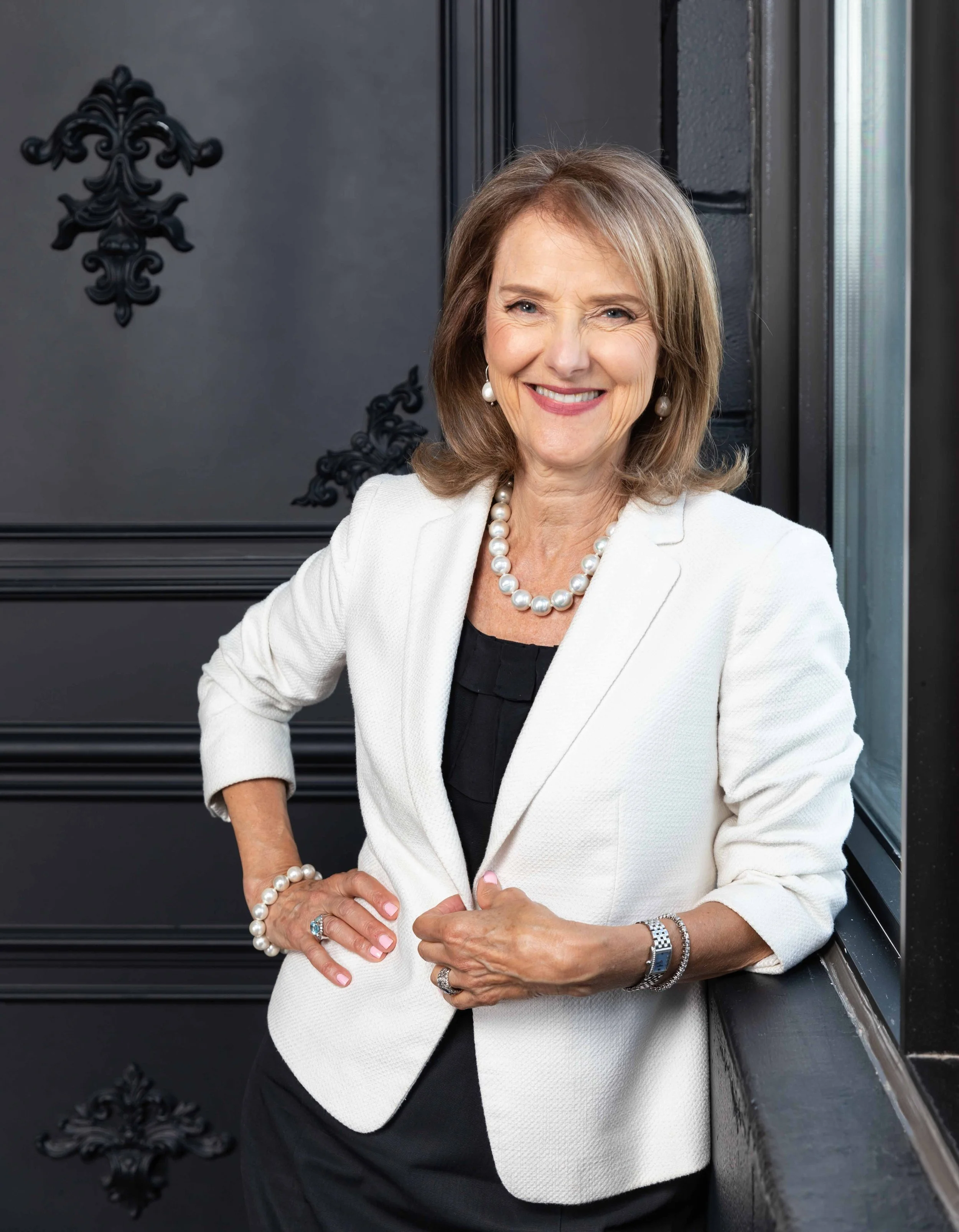 A middle-aged woman with shoulder-length light brown hair, smiling, dressed in a white blazer, black top, and pearl jewelry, standing near a window with black decorative wall accents in the background.