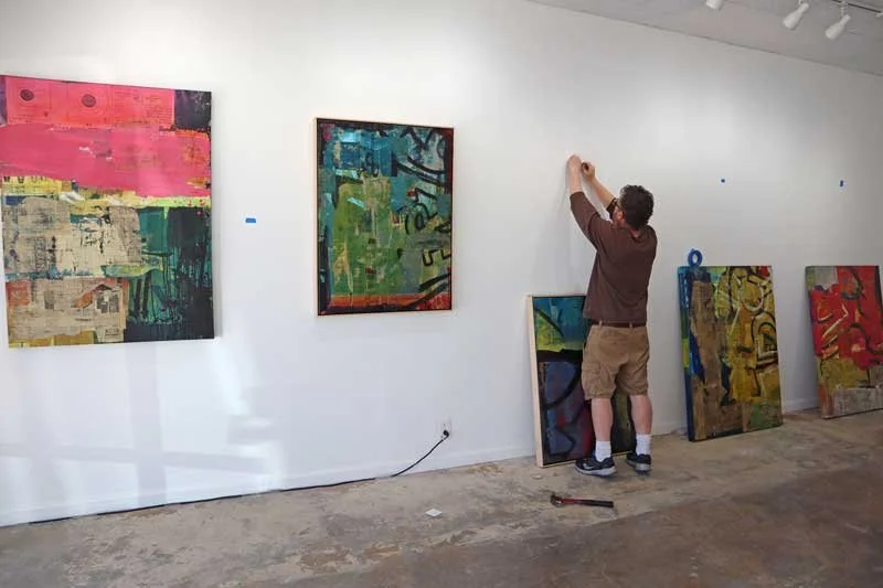 Man hanging artwork in art gallery with several colorful paintings leaning against the wall.