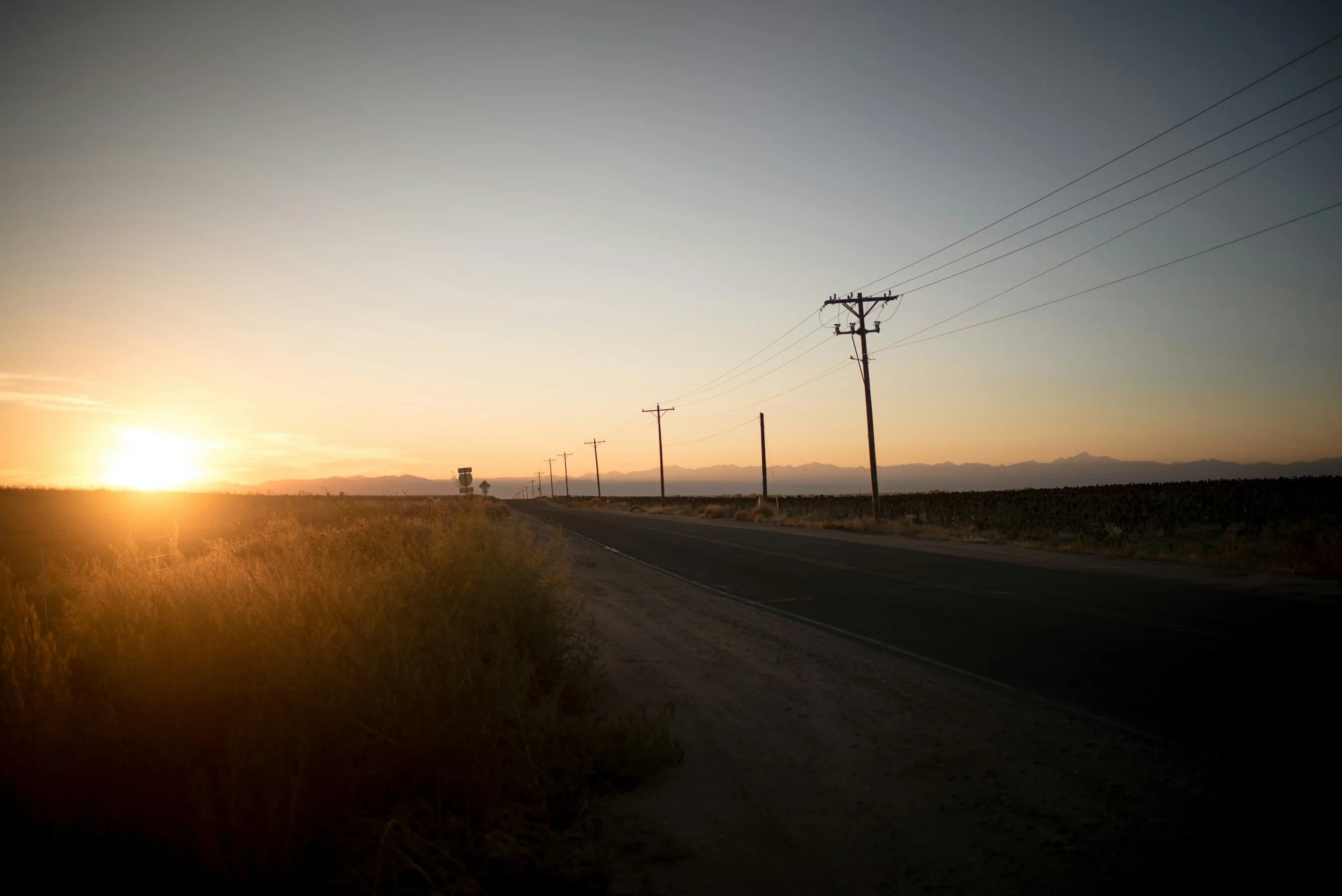 A rural road at sunset with power lines running along the side, open fields, and mountains in the distance.