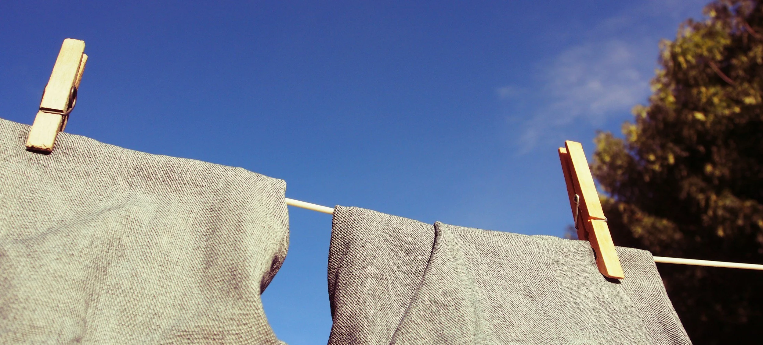 Gray fabric hanging on a clothesline outdoors, secured with wooden clothespins, against a clear blue sky and a background of trees.