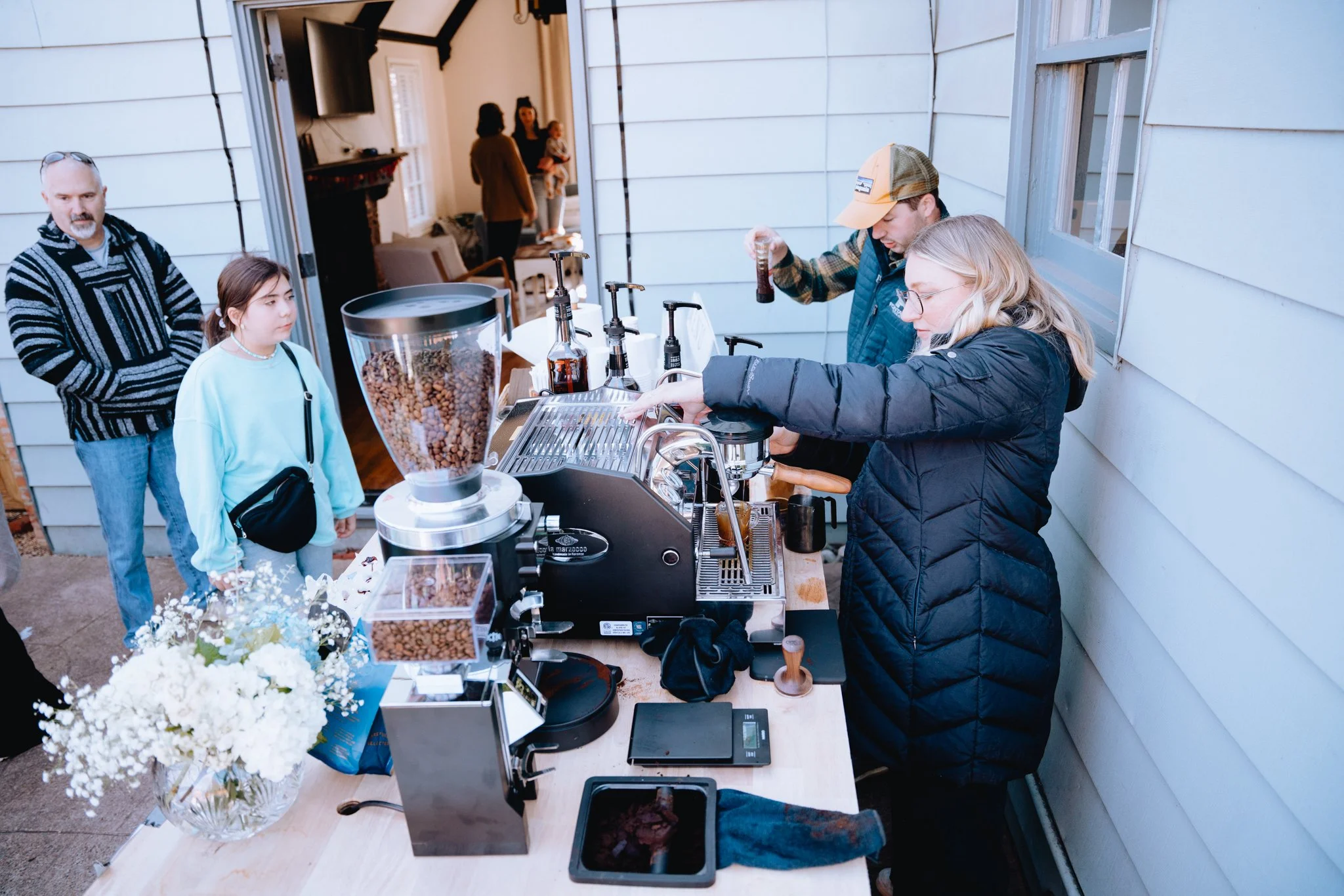 People serving coffee and food at an outdoor event table on a porch or patio, with a woman and a young girl in line and two workers behind the table preparing drinks.