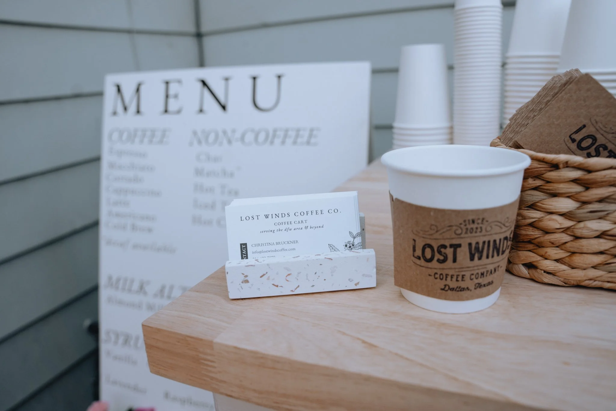Close-up of a coffee shop order station with a white paper menu, paper coffee cup with a cardboard sleeve, and a basket with brown napkins, on a wooden counter.