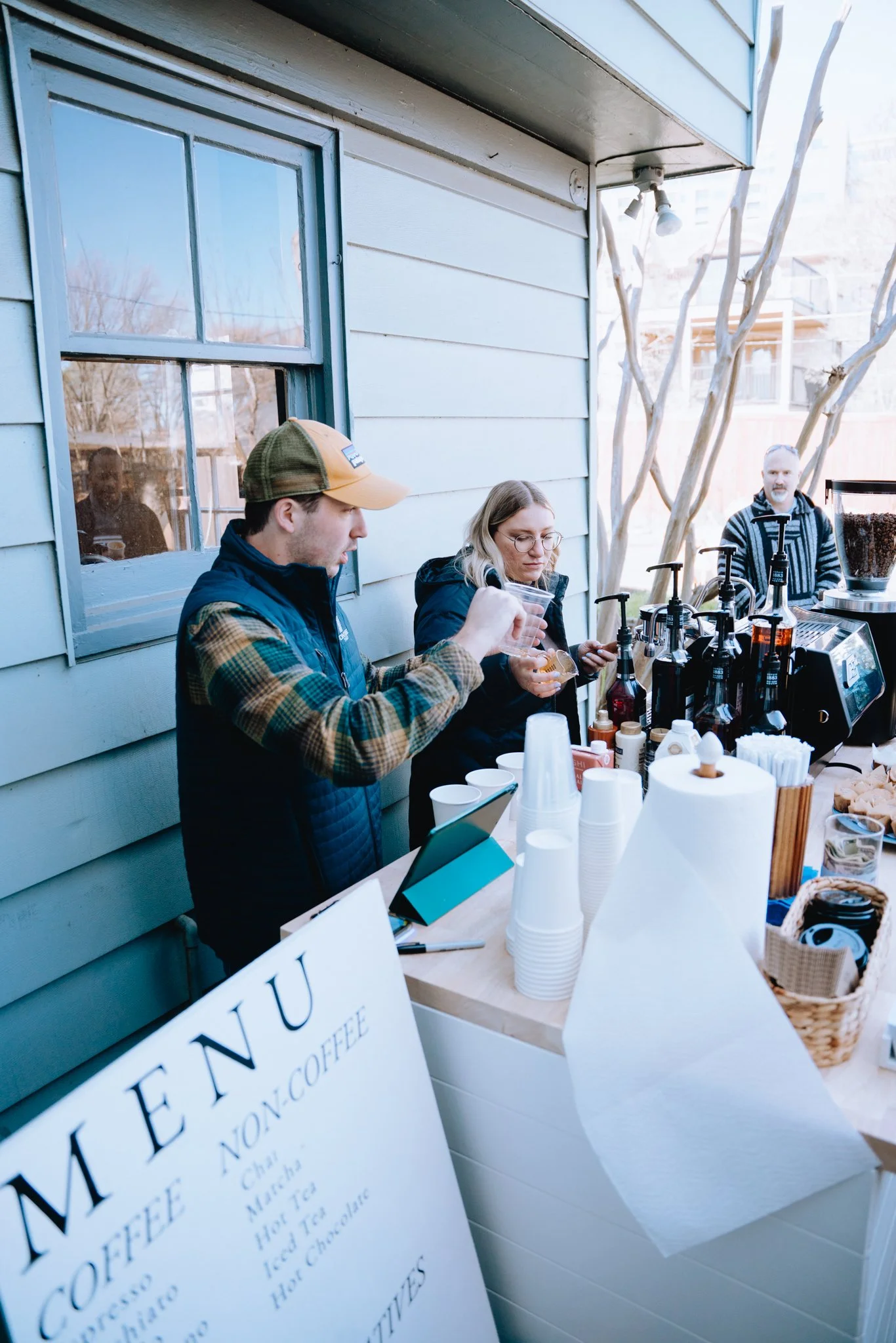 People ordering coffee at an outdoor coffee stand with various syrups and cups on the counter, and a sign advertising non-coffee drinks.