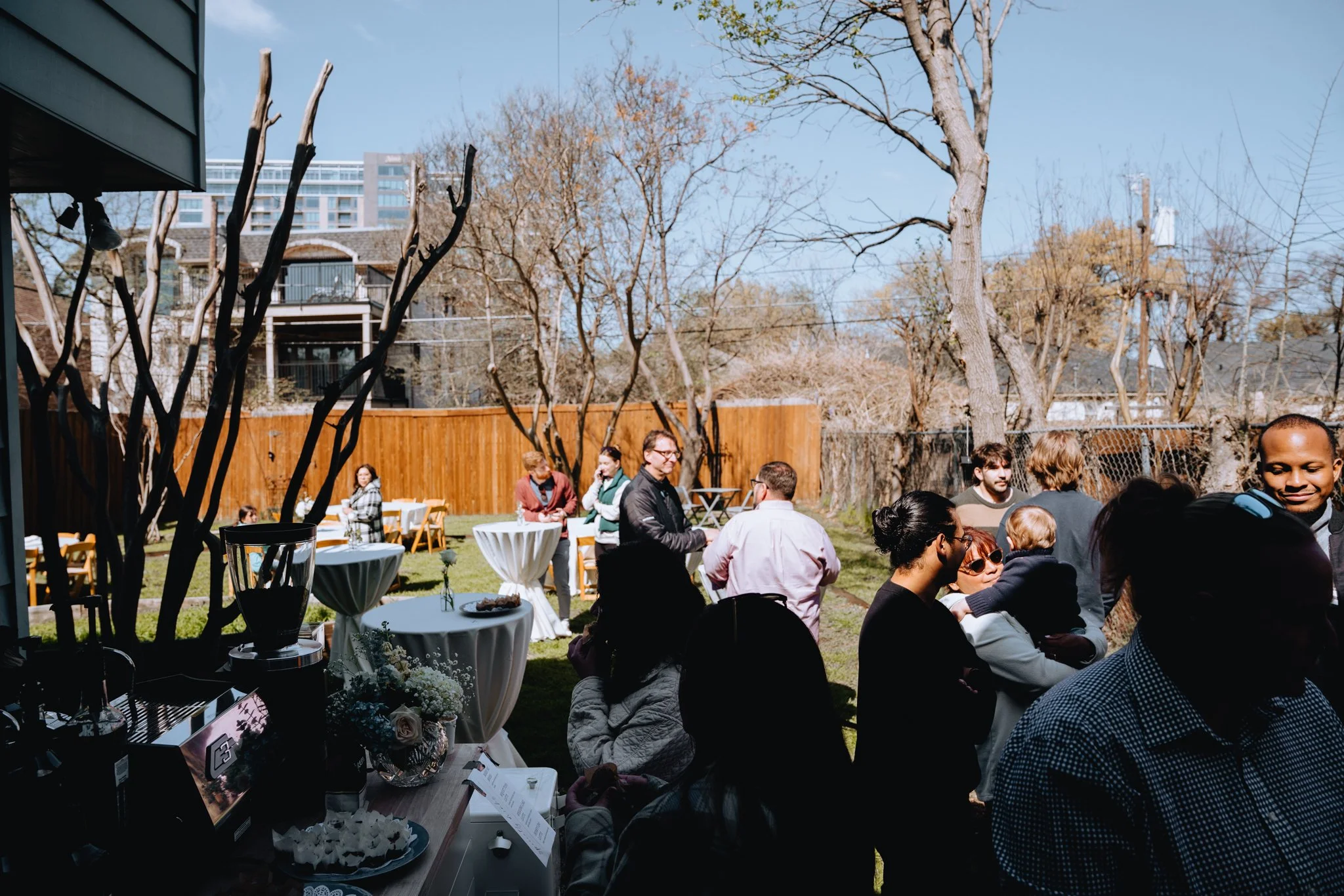 People gathered outdoors at a social event with tables and chairs, trees in the background, and a wooden fence, under a clear blue sky.