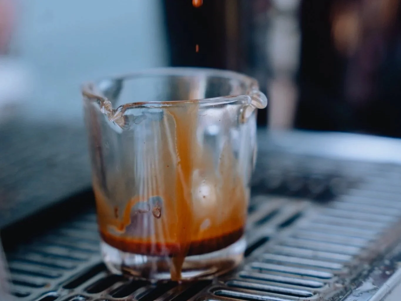 Close-up of a glass espresso shot sitting on a metal drip tray, with coffee dripping down the side of the glass.